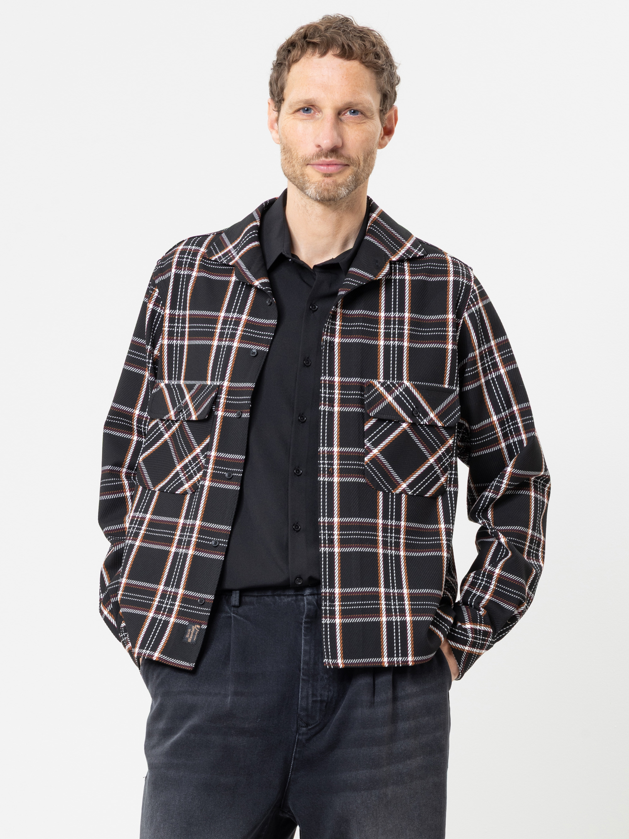 Ein Mann mit kurzen Locken trägt ein schwarzes Hemd mit Knöpfen und eine schwarz-weiß-rot karierte Jacke mit großen Vordertaschen und steht vor einem schlichten weißen Hintergrund.