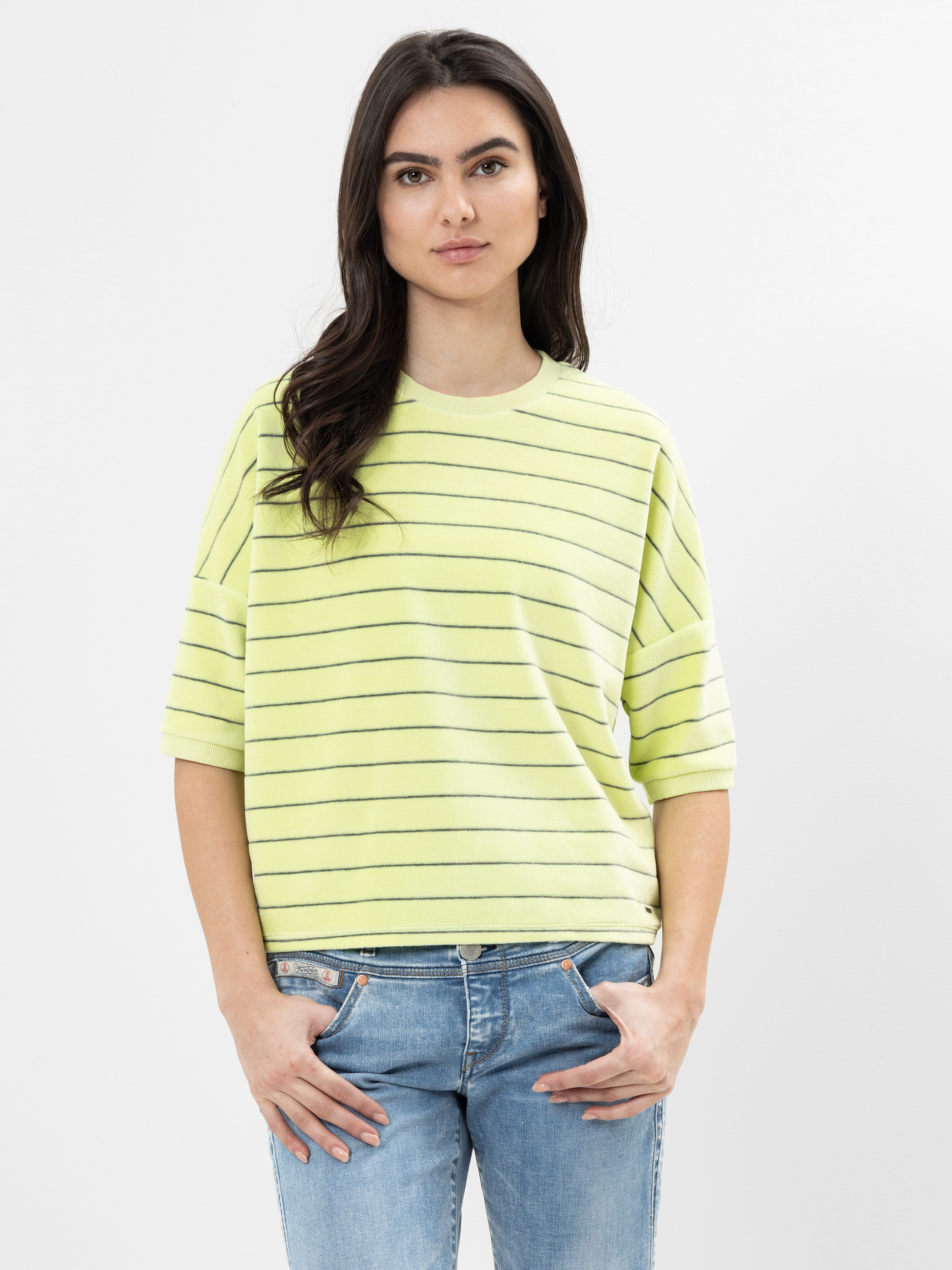 Eine Frau mit langen dunklen Haaren, die ein hellgelbes, kurzärmeliges, gestreiftes Hemd und blaue Jeans trägt, steht vor einem schlichten weißen Hintergrund und schaut mit neutralem Blick in die Kamera.
