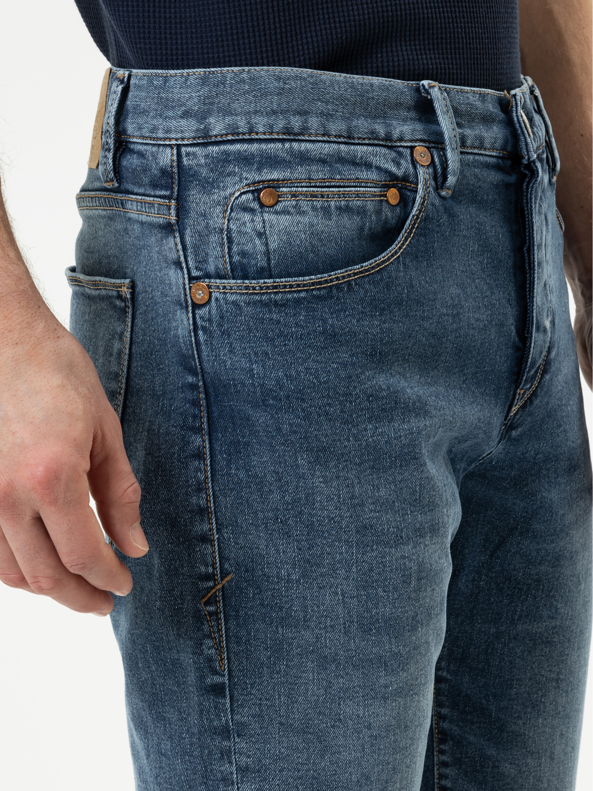 Nahaufnahme einer Person, die eine blaue Jeans und ein dunkles Hemd trägt. Zu sehen sind die Vordertasche, Gürtelschlaufen, Kupfernieten und ein Teil der linken Hand an der Seite.