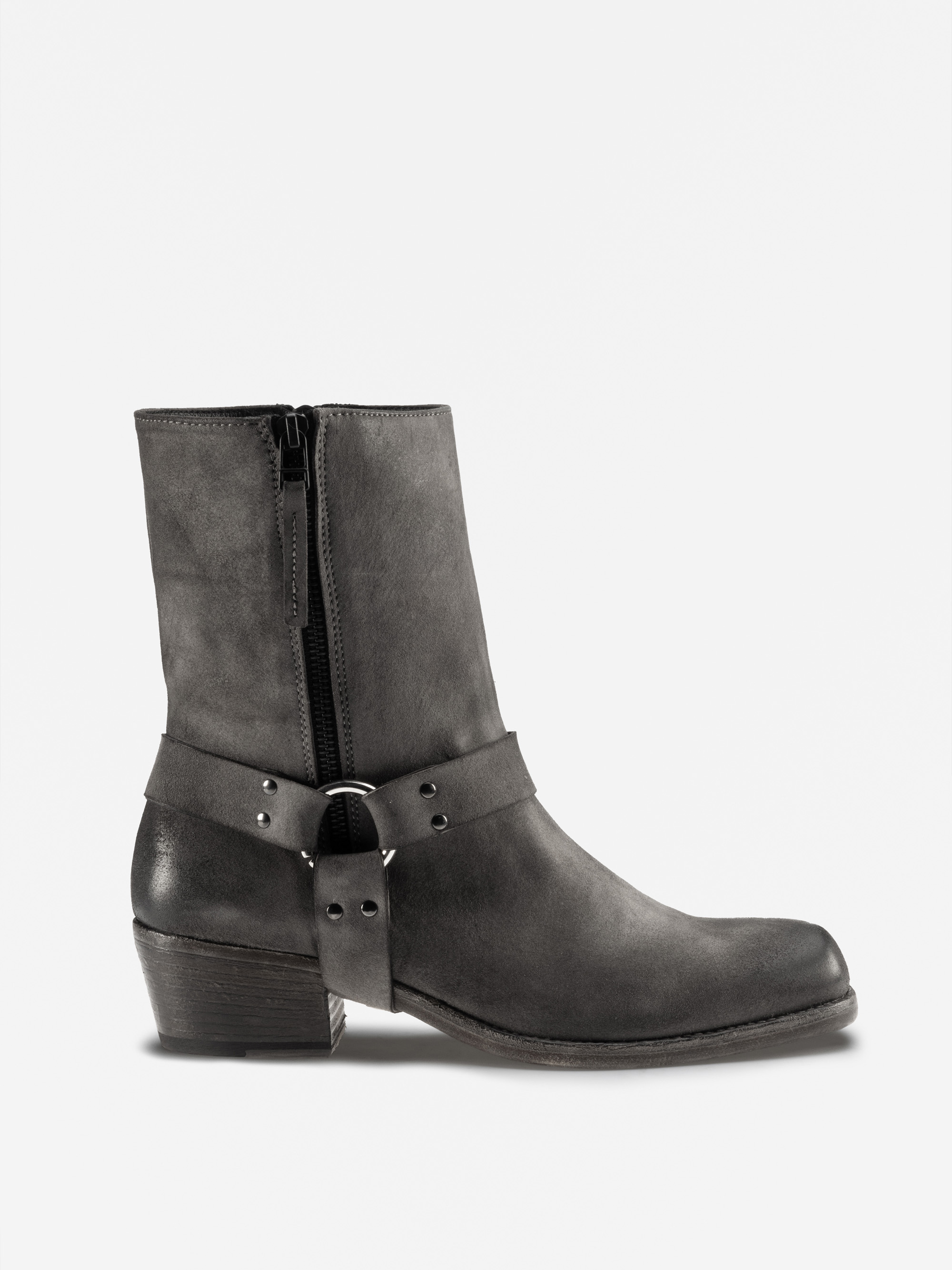 Eine schwarze Lederstiefelette mit niedrigem, klobigem Absatz, seitlichem Reißverschluss und dekorativem Riemen mit Metallnieten und Ringen um den Knöchel. Der Stiefel ist von der Seite vor einem weißen Hintergrund fotografiert.