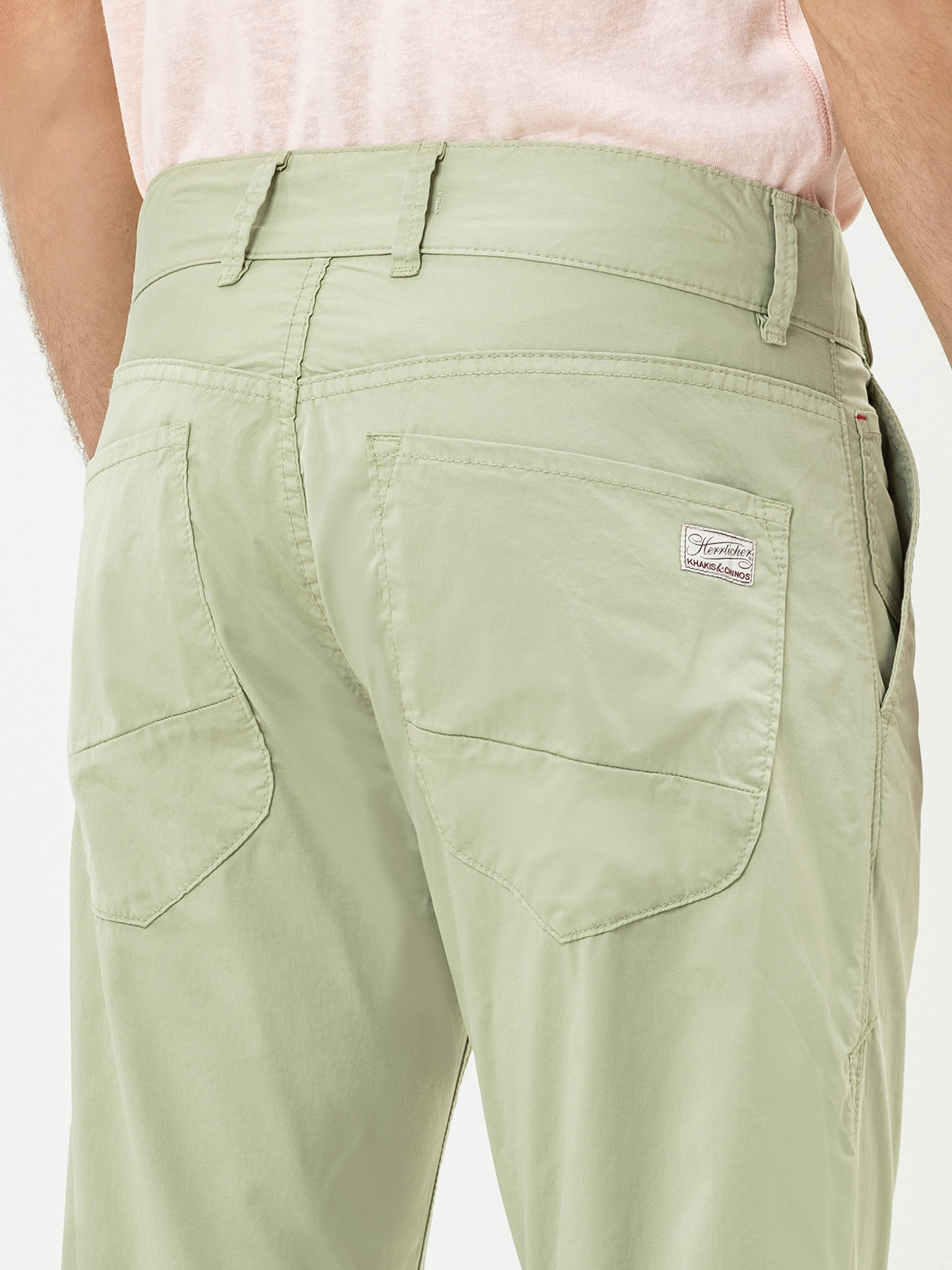 Eine Person, die eine hellgrüne Hose mit Gesäßtaschen und einem kleinen aufgenähten Etikett auf der rechten Tasche trägt; außerdem ist ein Teil eines hellrosa Hemdes zu sehen.