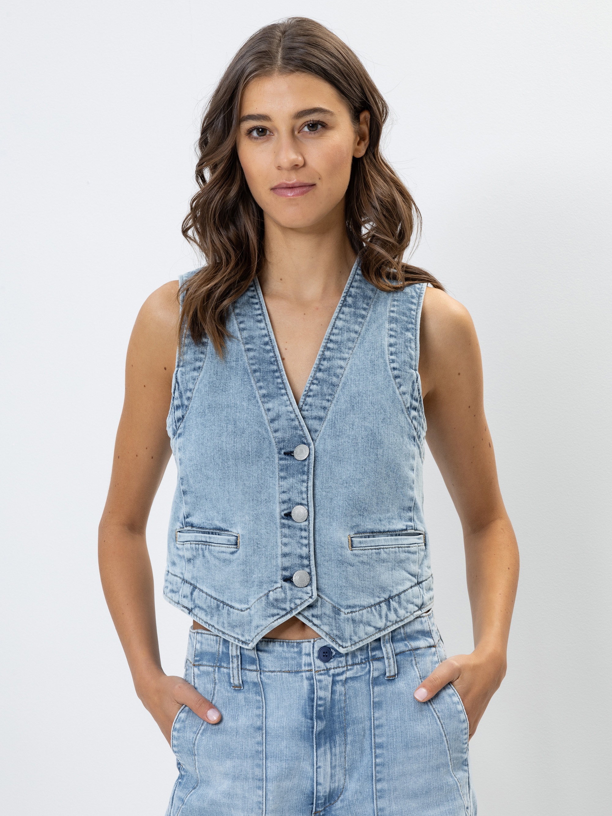 Eine Frau mit gewelltem braunem Haar trägt eine hellblaue Jeansweste und eine dazu passende Jeans mit hoher Taille. Sie steht vor einem schlichten weißen Hintergrund, hat die Hände in den Taschen und schaut in die Kamera.