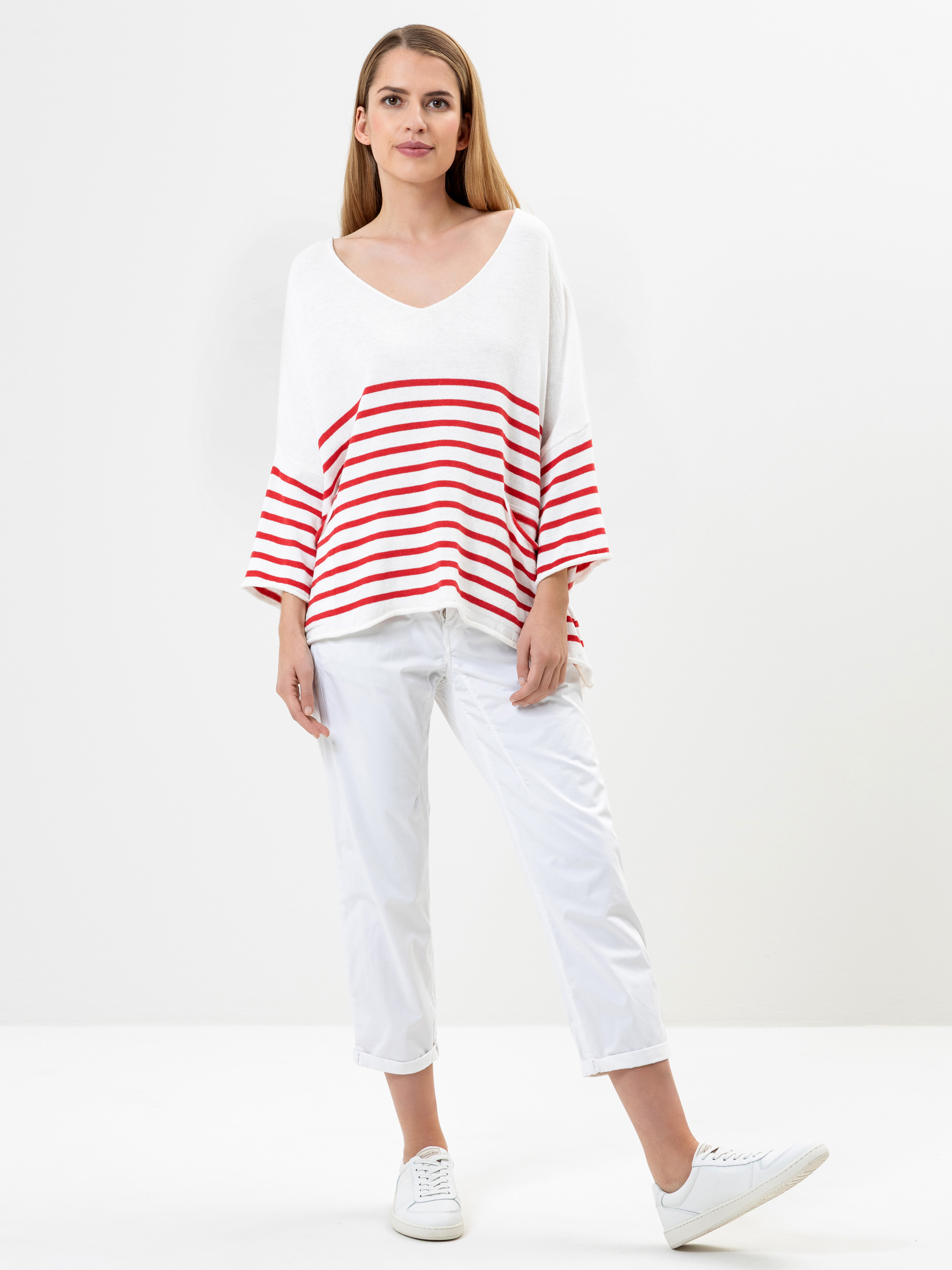 Eine Frau steht vor einem weißen Hintergrund und trägt ein lockeres weißes Oberteil mit roten Querstreifen und Dreiviertelärmeln, eine weiße kurze Hose und weiße Turnschuhe. Sie hat langes, glattes Haar und einen neutralen Gesichtsausdruck.