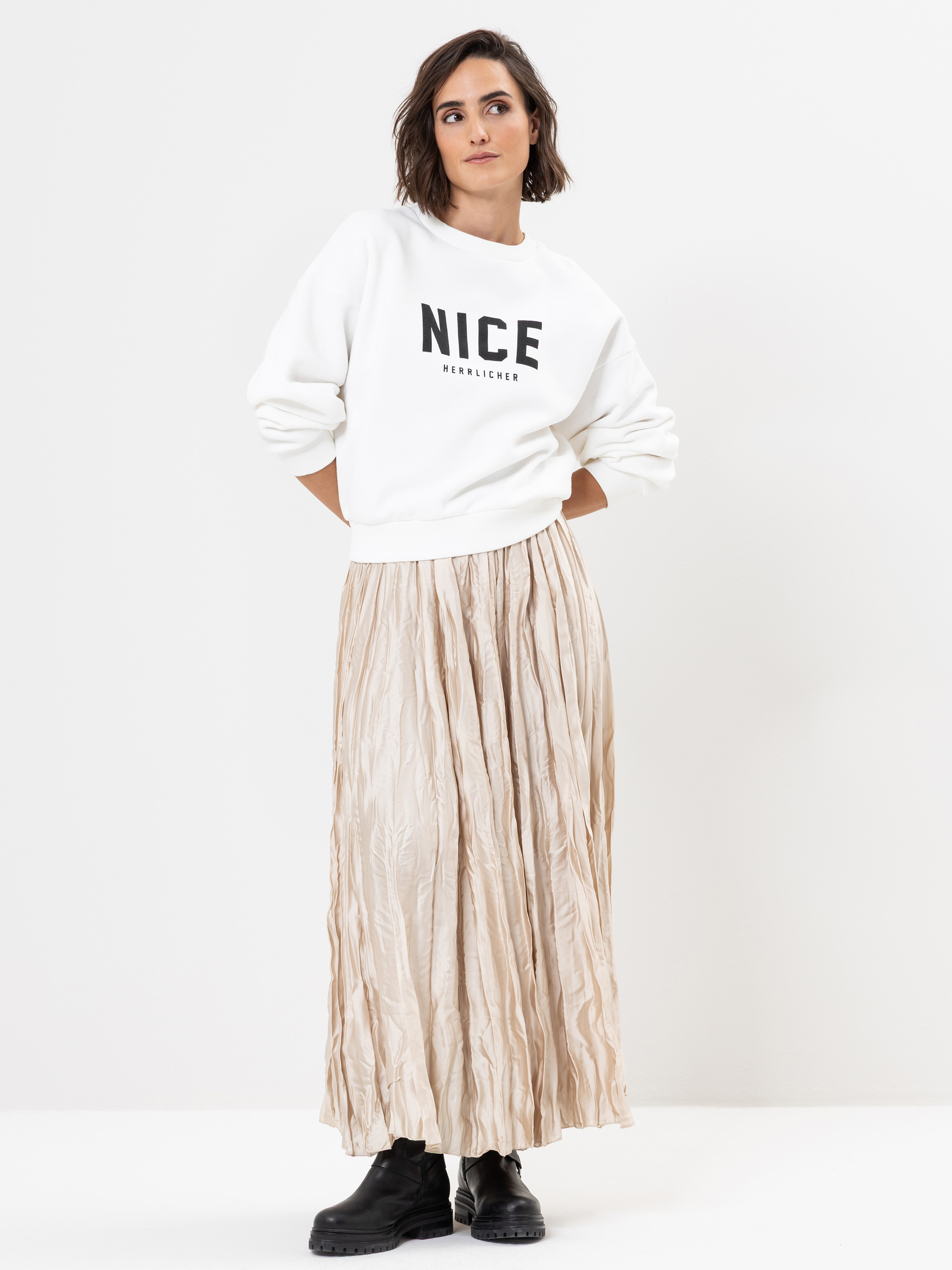 Eine Frau steht vor einem weißen Hintergrund und trägt ein weißes Sweatshirt mit dem Aufdruck NICE, einen langen beigen Faltenrock und schwarze Stiefel. Sie blickt leicht zur Seite und hat einen entspannten Gesichtsausdruck.