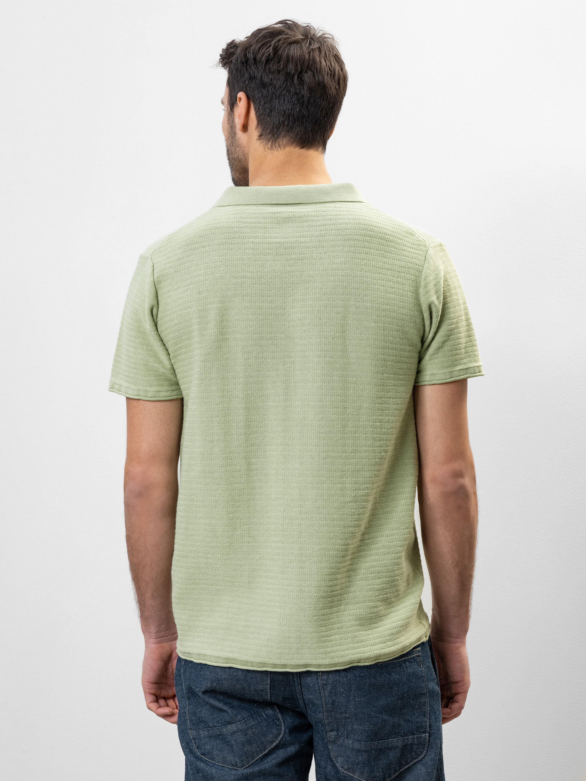 Ein Mann steht mit einem hellgrünen, kurzärmeligen Polohemd und einer dunkelblauen Jeans vor einem schlichten weißen Hintergrund und blickt von der Kamera weg.
