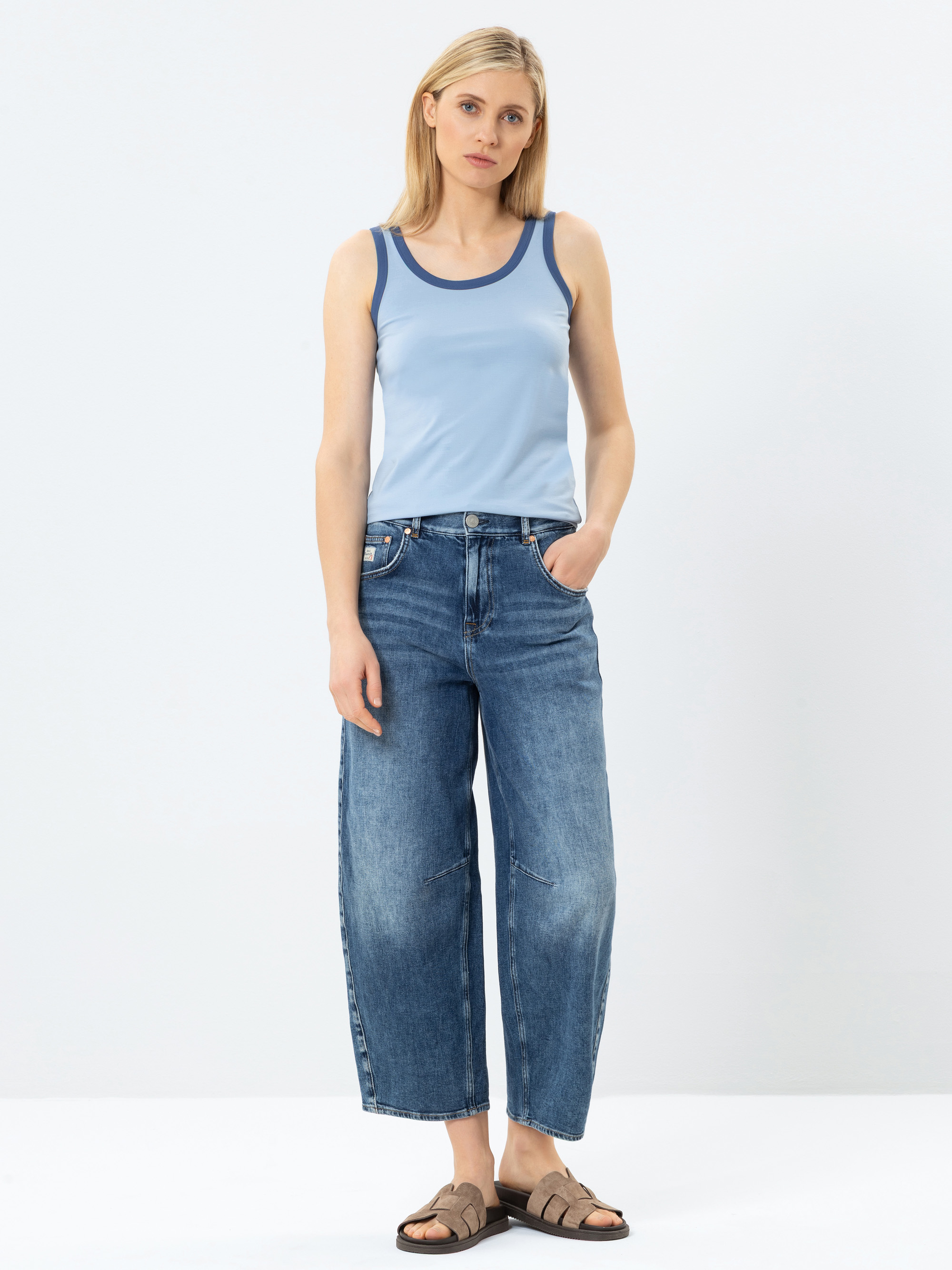 Eine Frau mit glattem blondem Haar trägt ein hellblaues Tanktop, locker sitzende Bluejeans und braune Sandalen. Sie steht vor einem schlichten weißen Hintergrund und hat eine Hand in ihrer Tasche.