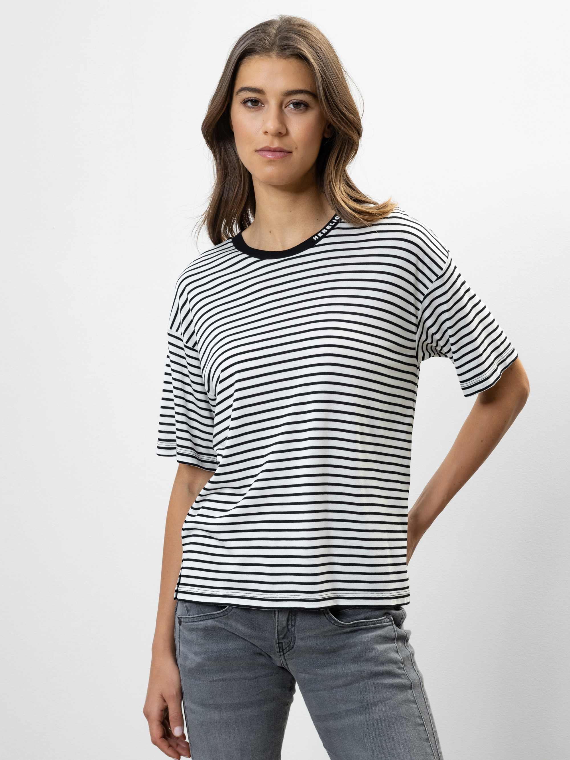 Eine Frau steht vor einem schlichten weißen Hintergrund und trägt ein schwarz-weiß gestreiftes kurzärmeliges Hemd und graue Jeans. Sie hat langes braunes Haar und einen neutralen Gesichtsausdruck, wobei sie eine Hand auf dem Rücken verschränkt hat.