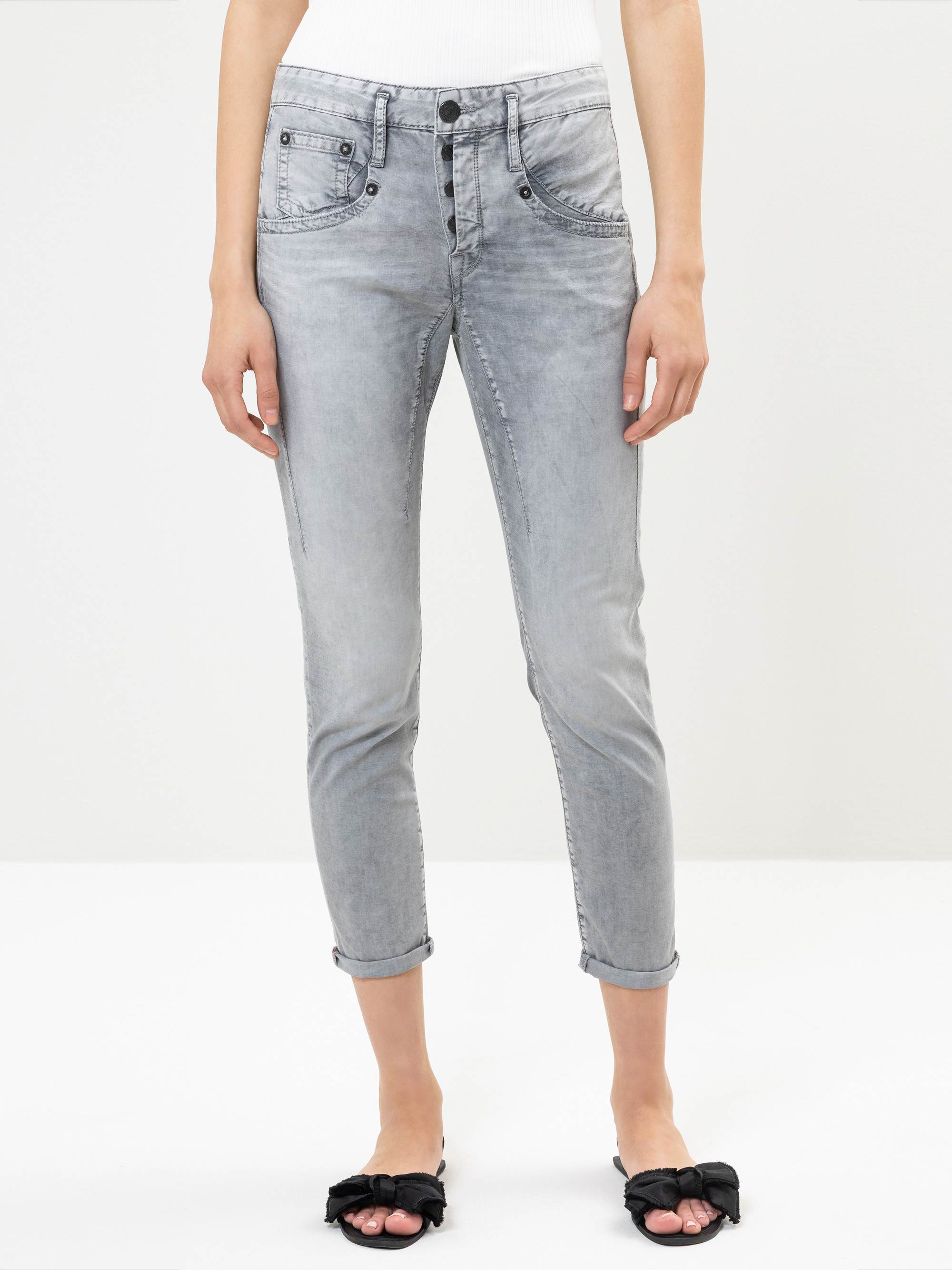 Eine Person, die hellgraue, hochgekrempelte Jeans mit Knopfleiste, ein weißes geripptes Tanktop und schwarze offene Sandalen mit Schleifen trägt, steht vor einem schlichten weißen Hintergrund.