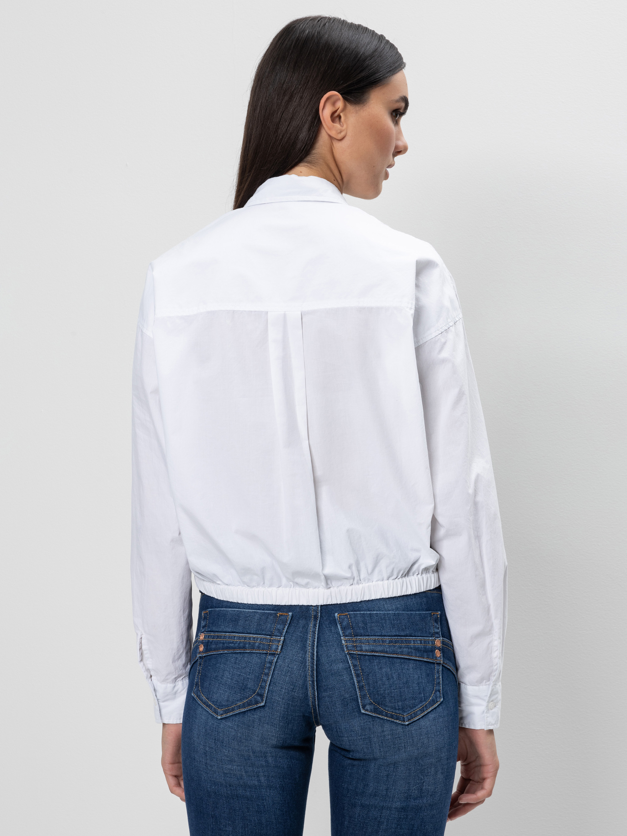 Eine Frau mit langen dunklen Haaren, die ein weißes langärmeliges Hemd und blaue Jeans trägt, steht mit dem Rücken zur Kamera vor einem schlichten hellen Hintergrund.