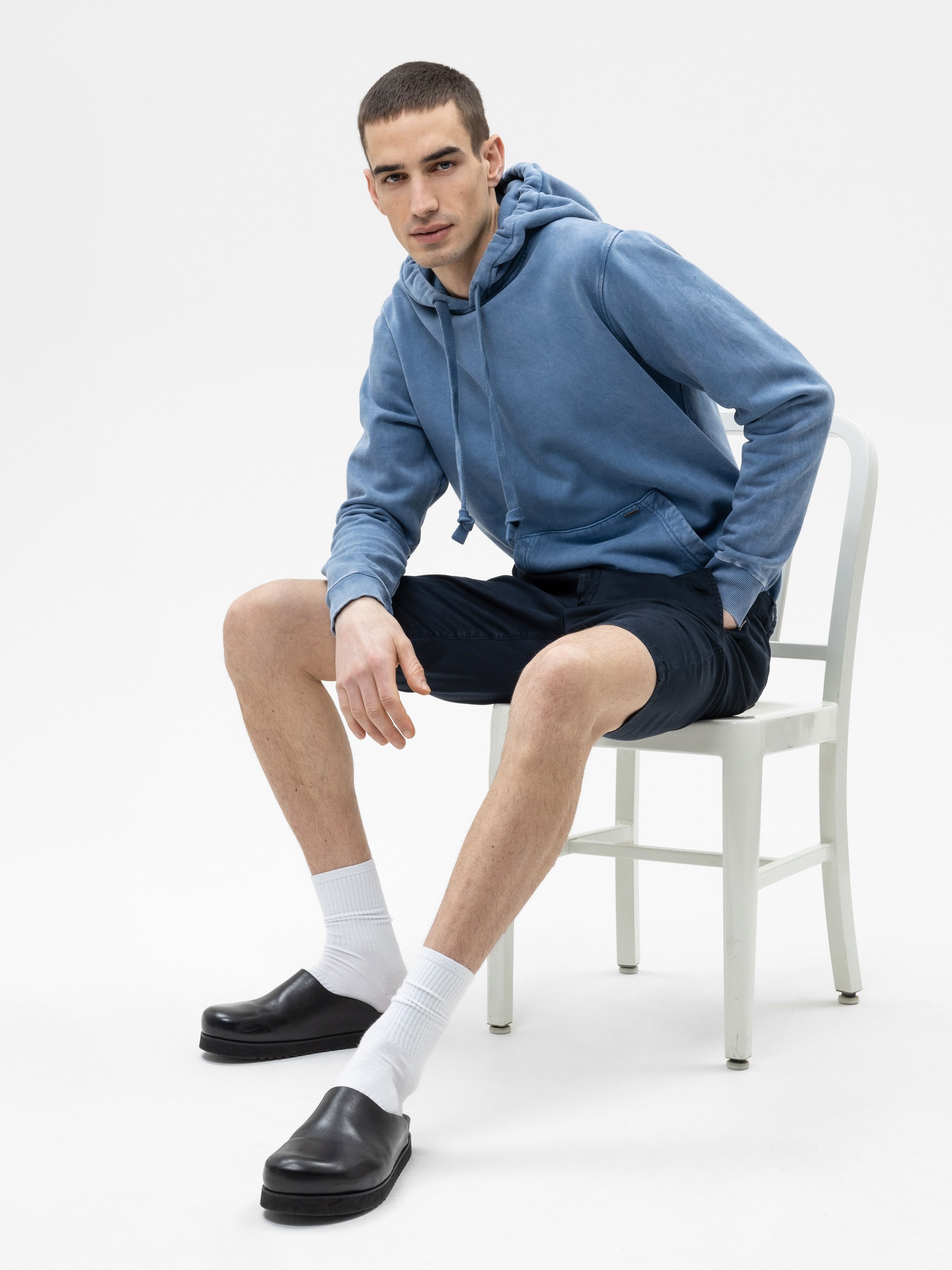 Ein Mann, der einen blauen Kapuzenpullover, marineblaue Shorts, weiße Socken und schwarze Slipper trägt, sitzt auf einem weißen Stuhl vor einem schlichten weißen Hintergrund.