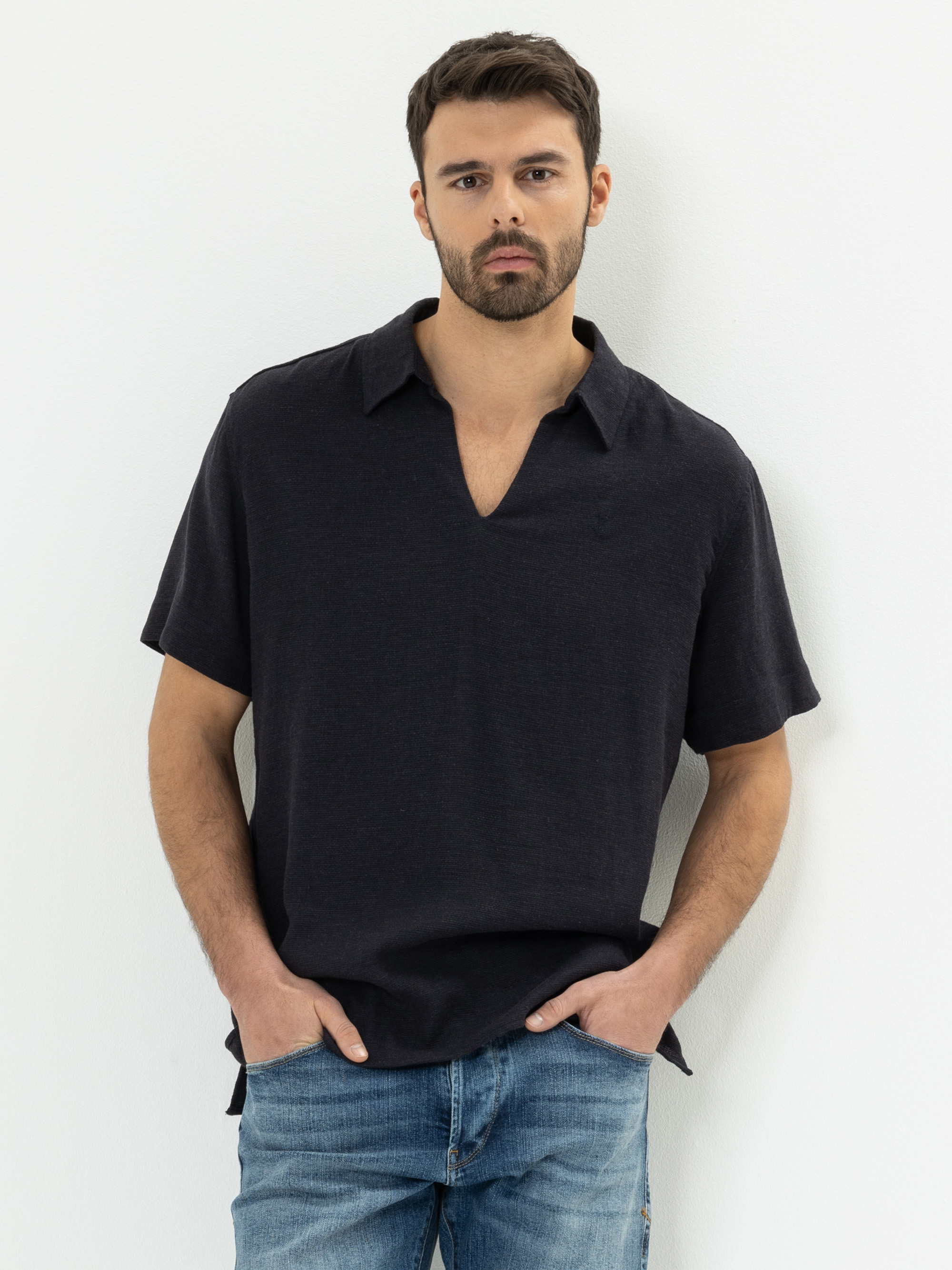 Ein Mann mit kurzen dunklen Haaren und Bart steht vor einer weißen Wand. Er trägt ein schwarzes kurzärmeliges Kragenhemd mit V-Ausschnitt und blaue Jeans. Er hat die Hände in den Taschen und blickt direkt in die Kamera.