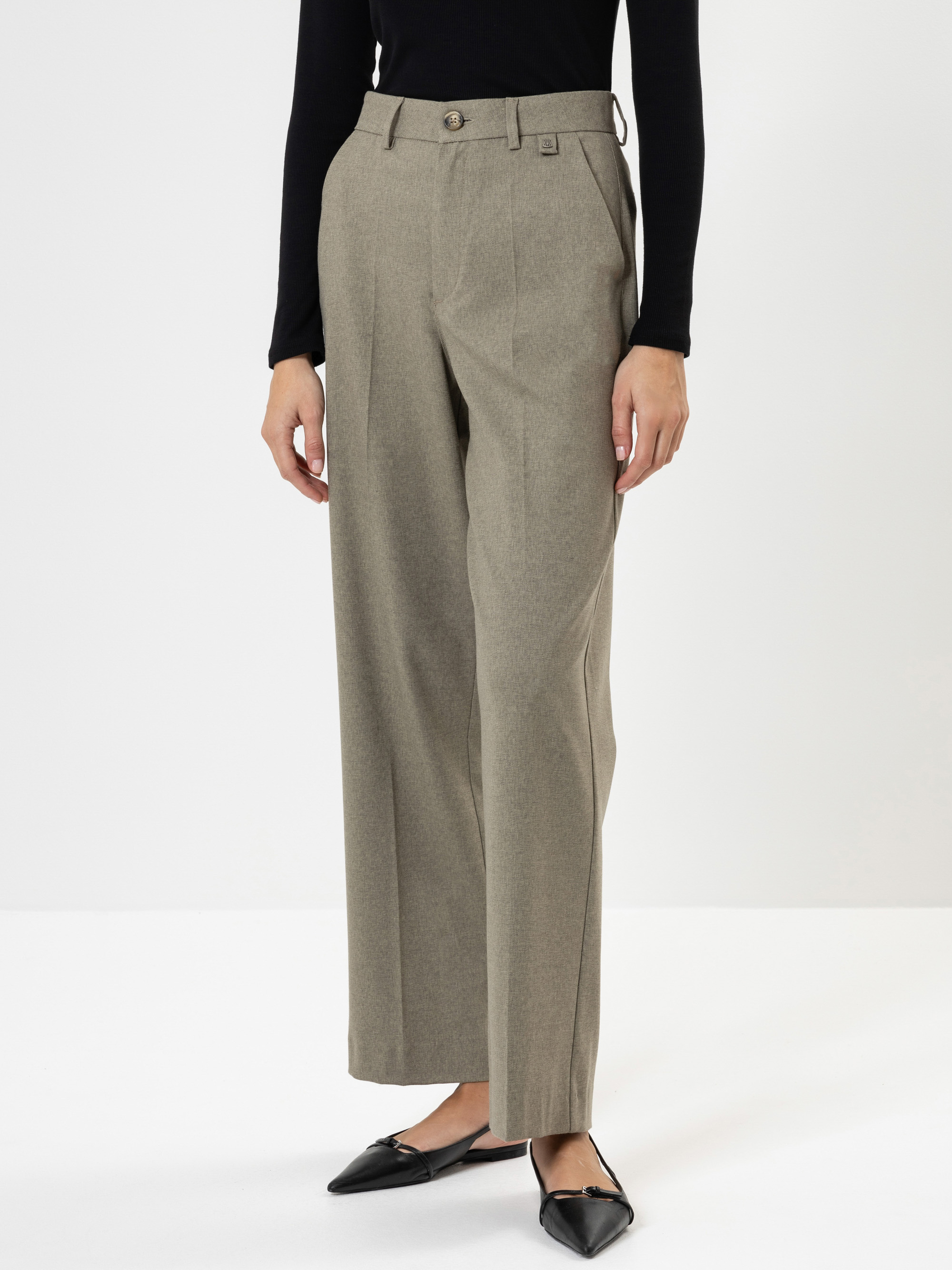 Eine Person, die eine hellgraue Anzughose mit hoher Taille und weitem Bein, ein schwarzes langärmeliges Oberteil und schwarze flache Schuhe mit spitzen Zehen trägt, steht vor einem schlichten weißen Hintergrund.