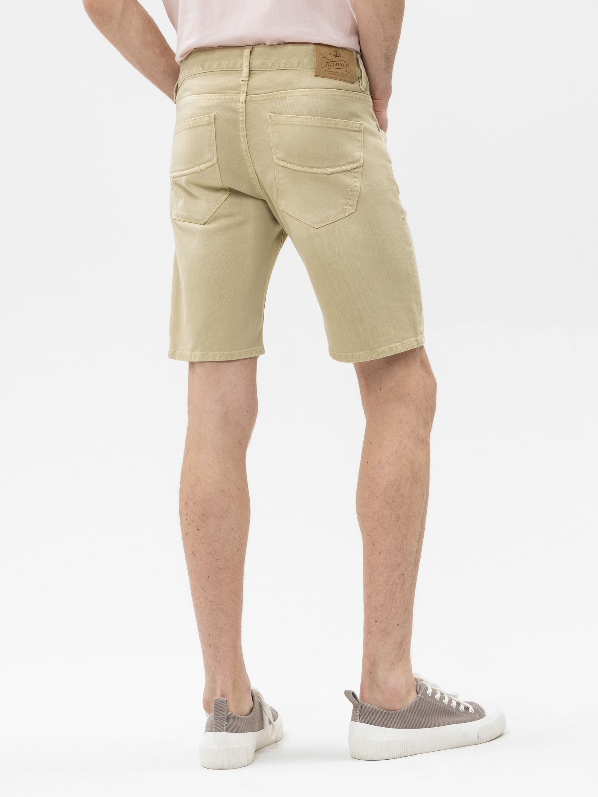 Eine Person, die hellbeige Shorts, ein hellrosa Hemd und grau-weiße Turnschuhe trägt, steht mit dem Rücken zur Kamera vor einem weißen Hintergrund.