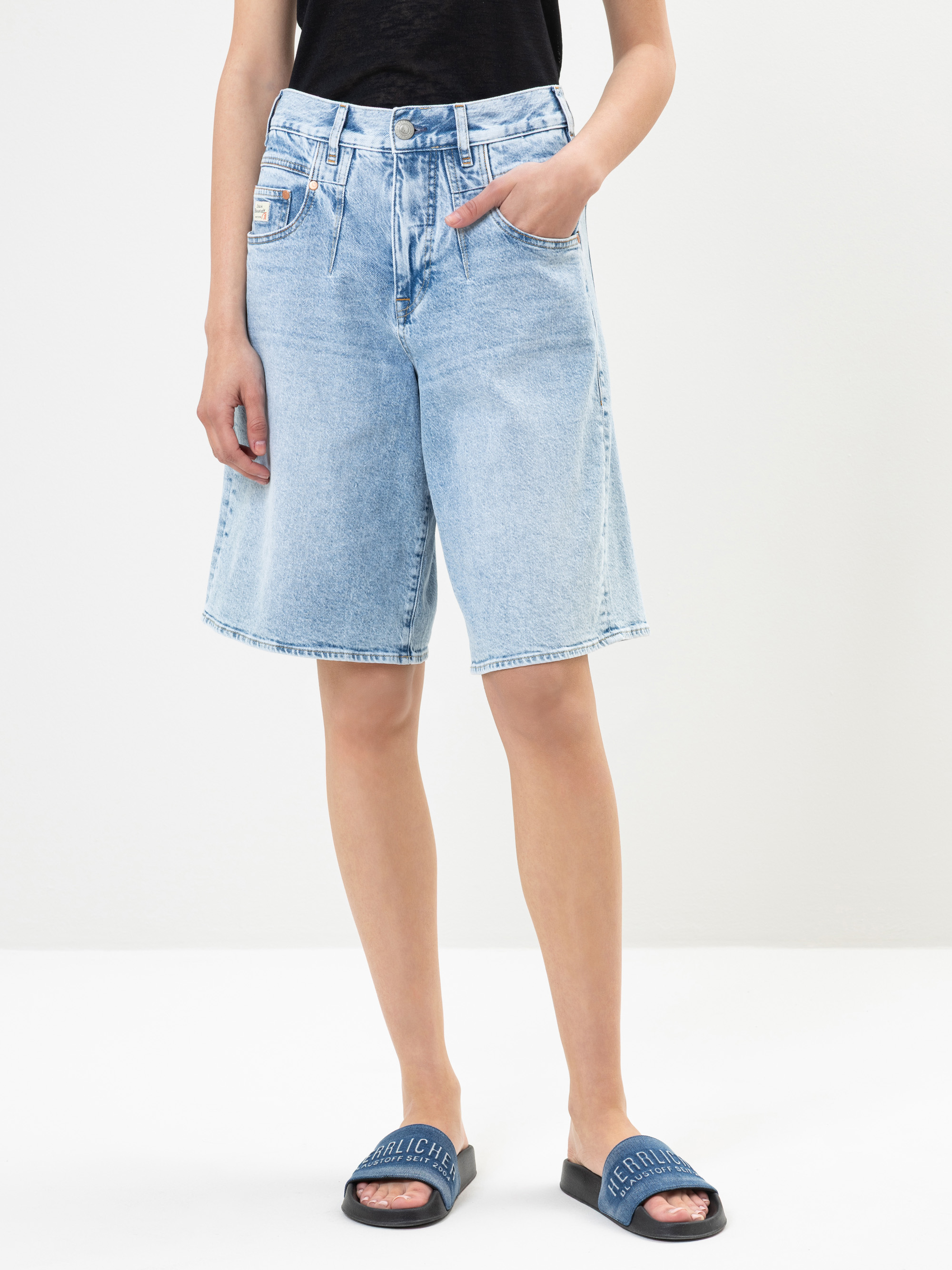 Eine Person, die hellblaue, knielange Jeans-Shorts, ein schwarzes Oberteil und blaue Slide-Sandalen trägt, steht vor einem schlichten weißen Hintergrund und hat eine Hand in der Tasche.