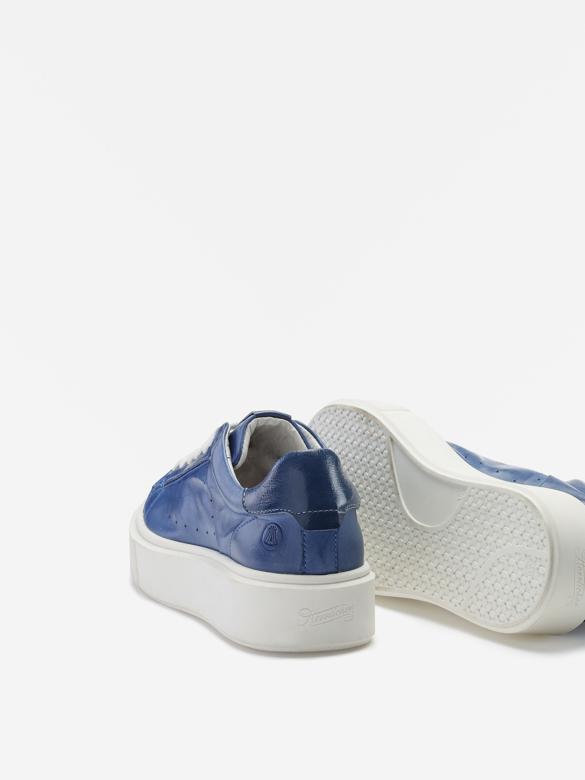 Ein Paar blaue Lederturnschuhe mit dicken weißen Sohlen; ein Schuh steht aufrecht und zeigt die Rückseite, während der andere auf der Seite liegt und die strukturierte Sohle zeigt.
