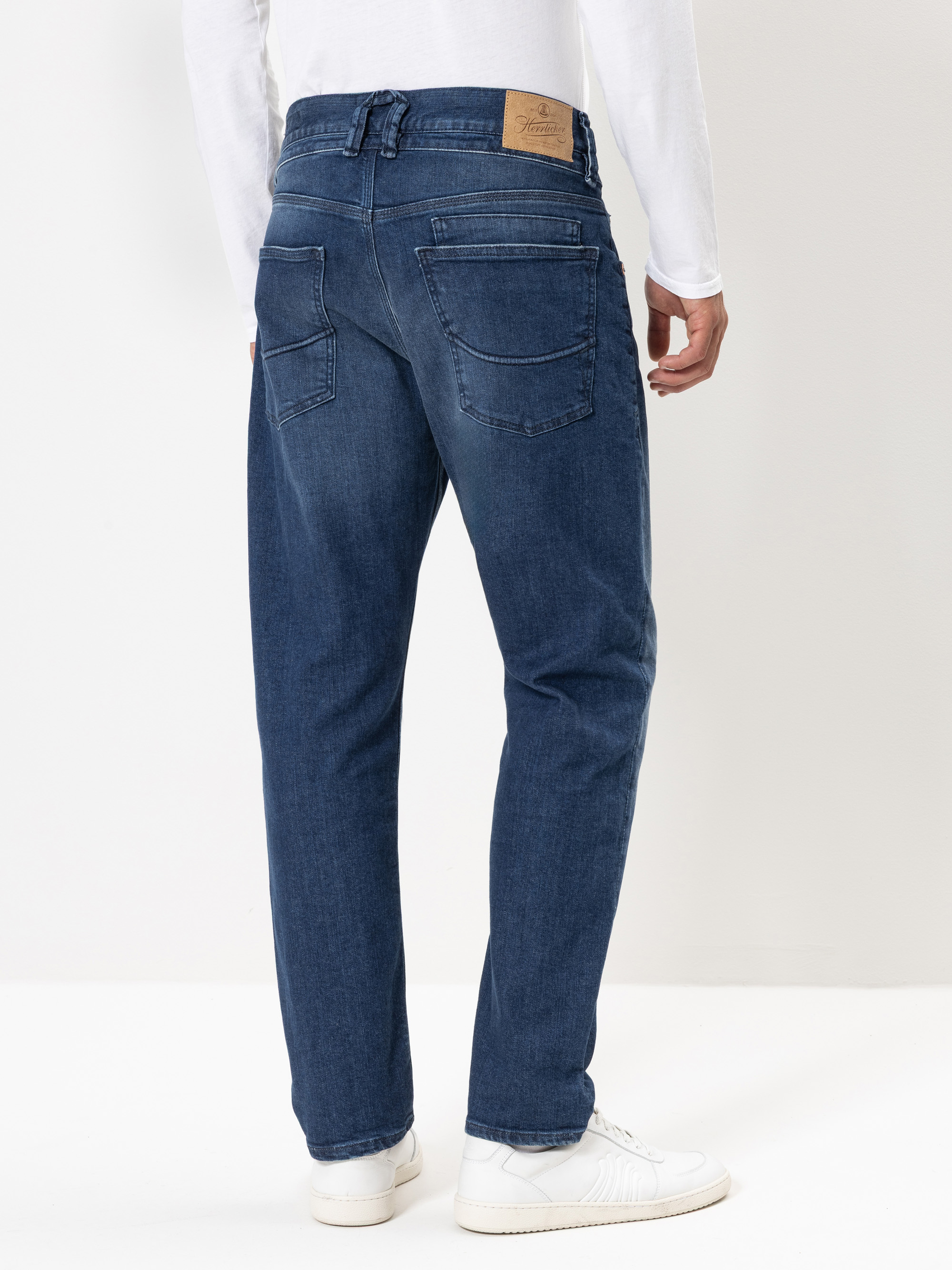 Eine Person, die eine blaue Jeans, ein weißes langärmeliges Hemd und weiße Turnschuhe trägt, steht mit dem Gesicht nach hinten und zeigt die Rückenansicht der Jeans vor einem schlichten hellen Hintergrund.