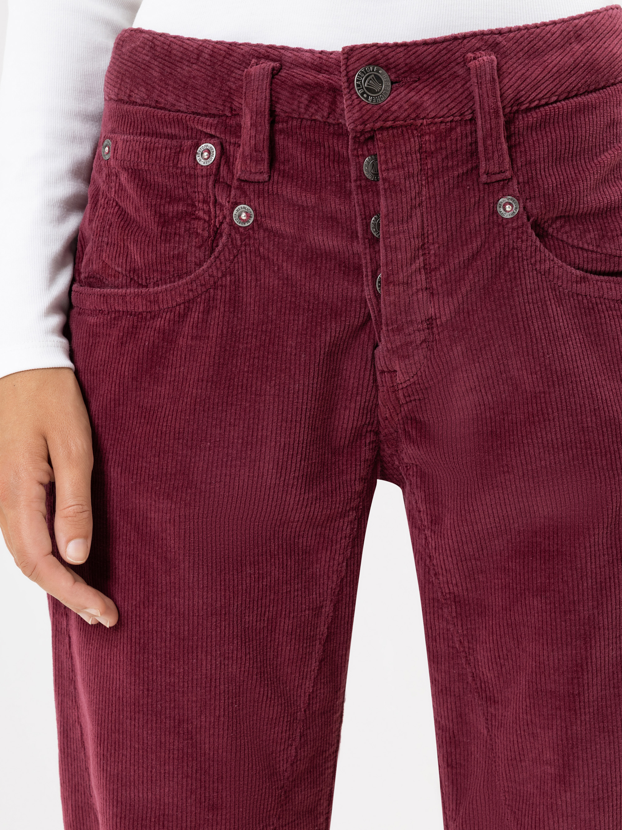 Eine Person trägt eine hochtaillierte, dunkelrote Cordhose mit mehreren Knöpfen an der Vorderseite und ein weißes langärmeliges Oberteil. Das Bild zeigt die Hose von der Taille bis zur Mitte des Oberschenkels, wobei eine Hand sichtbar ist.