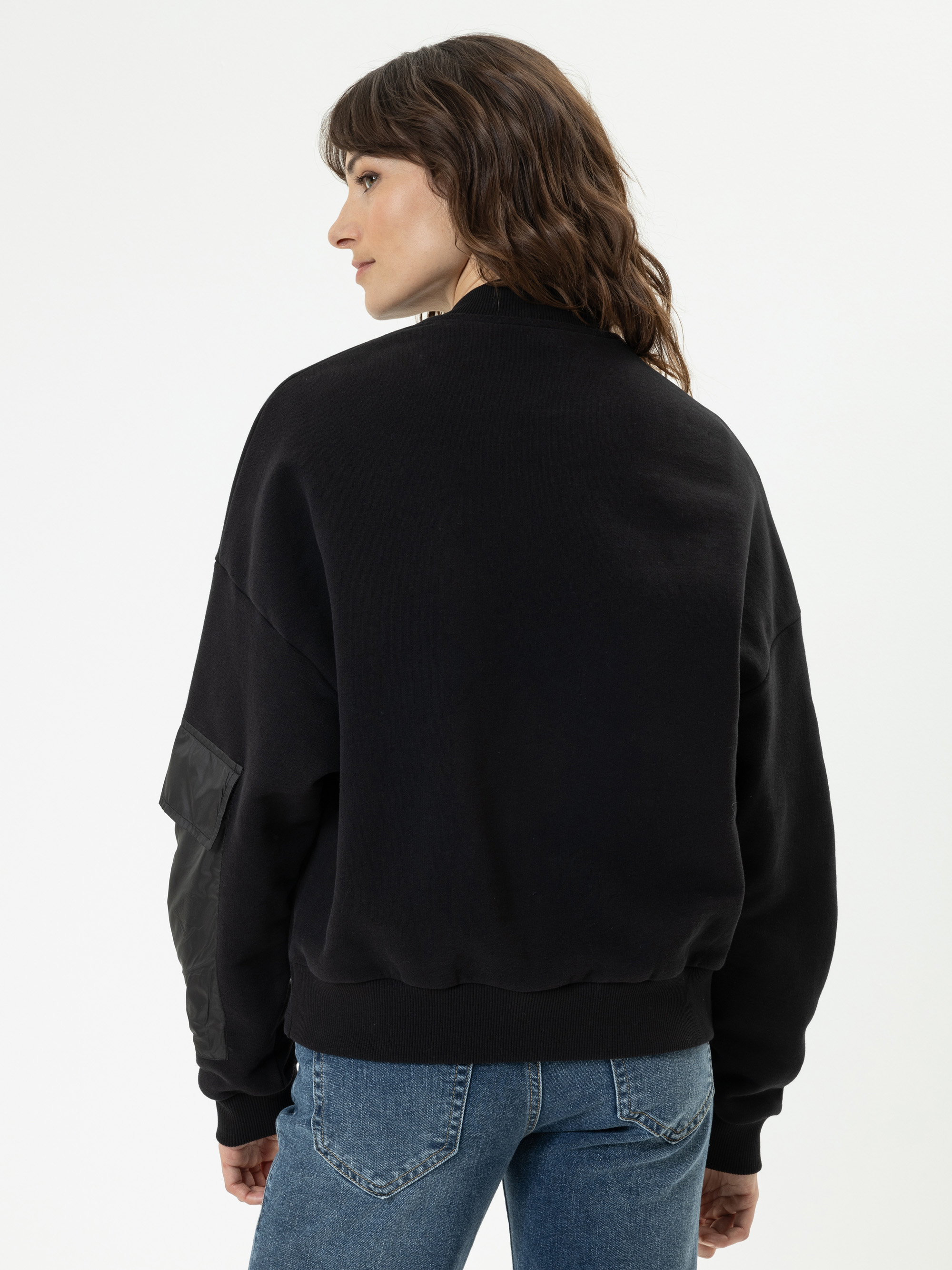 Eine Person mit gewelltem braunem Haar trägt ein schwarzes Sweatshirt mit einem Aufnäher am linken Ärmel und eine blaue Jeans. Sie steht vor einem einfarbigen weißen Hintergrund und blickt von der Kamera weg.