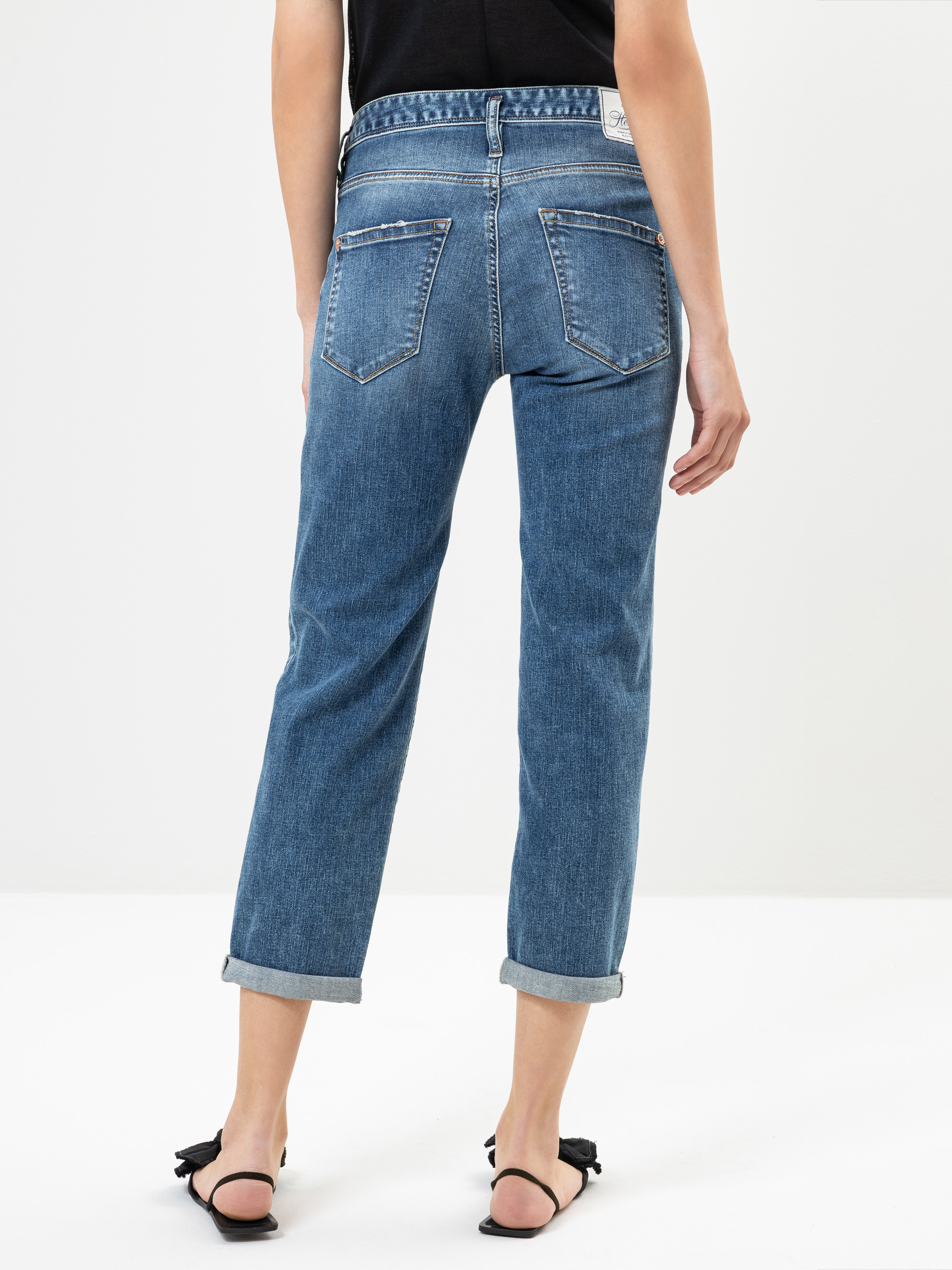 Eine Person trägt eine blaue Jeans mit Bündchen und schwarze hochhackige Sandalen. Das Foto ist von hinten aufgenommen und zeigt die Gesäßtaschen und die aufgerollten Säume der Jeans vor einem schlichten weißen Hintergrund.