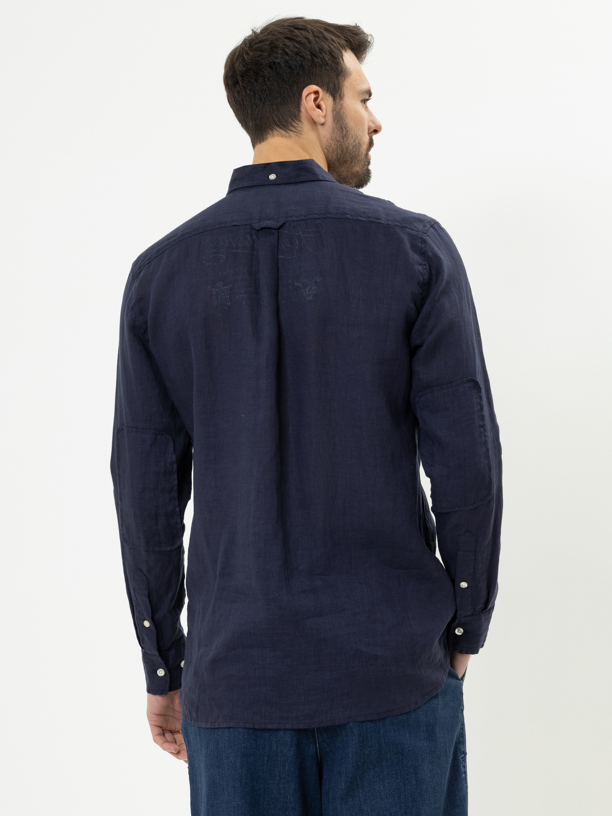 Ein Mann mit kurzen braunen Haaren ist von hinten zu sehen. Er trägt ein langärmeliges marineblaues Button-up-Hemd und blaue Jeans und steht vor einem schlichten weißen Hintergrund.