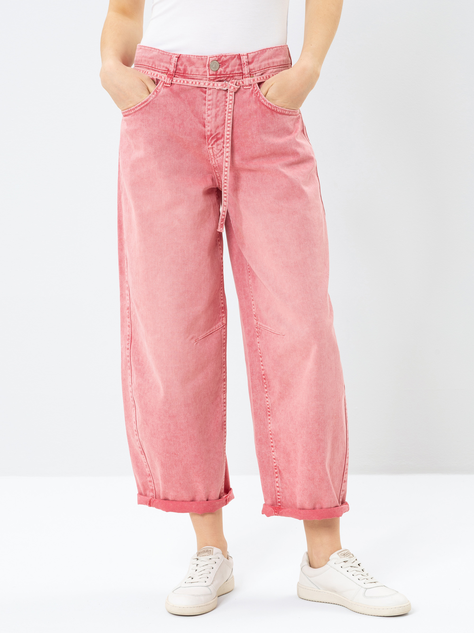 Person, die eine pinkfarbene Jeans mit hoher Taille und weitem Bein sowie einen passenden Gürtel, ein weißes Oberteil und weiße Turnschuhe trägt. Die Jeans sind an den Knöcheln mit Bündchen versehen, und die Person hat die Hände in den Taschen. Der