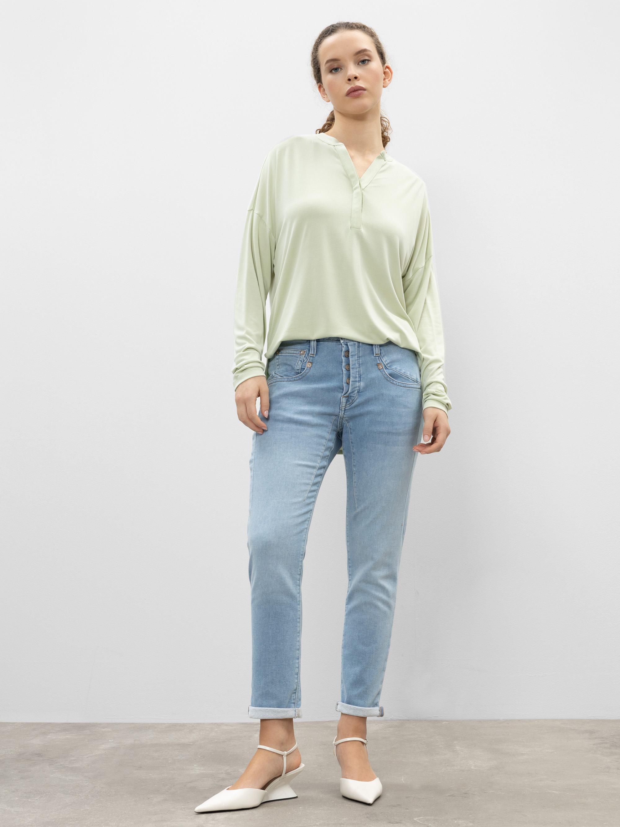 Eine Frau steht vor einer schlichten weißen Wand. Sie trägt eine hellgrüne, langärmelige Bluse, hellblaue Jeans mit gekrempelten Manschetten und weiße, spitz zulaufende Schuhe. Sie hat ihr lockiges Haar zurückgesteckt und nimmt eine entspannte, selb