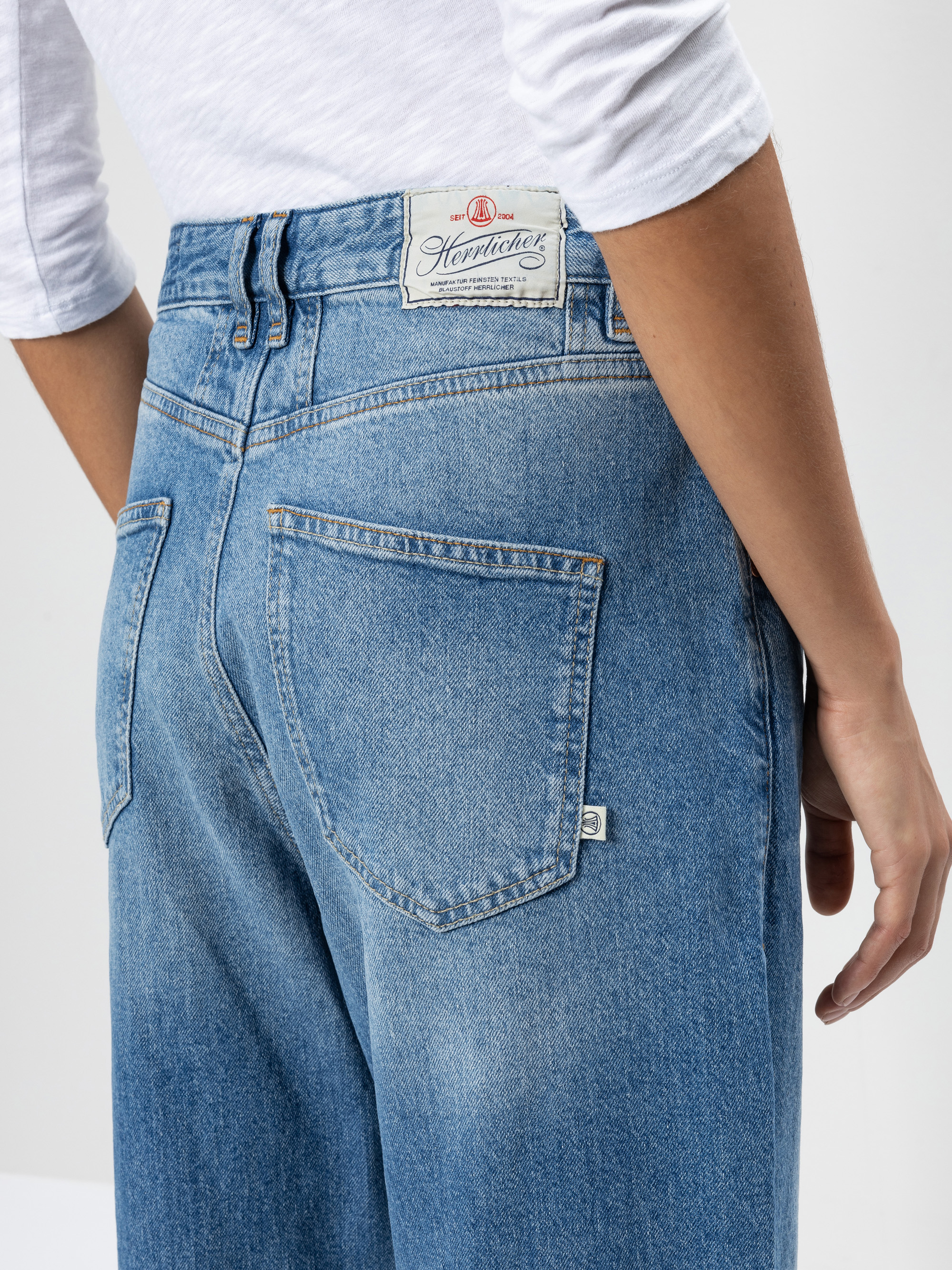 Eine Person, die hellblaue Jeans mit hoher Taille und ein weißes langärmeliges Hemd trägt, wird von hinten gezeigt. Die Jeans hat ein rechteckiges Etikett am Bund und zwei große Gesäßtaschen.