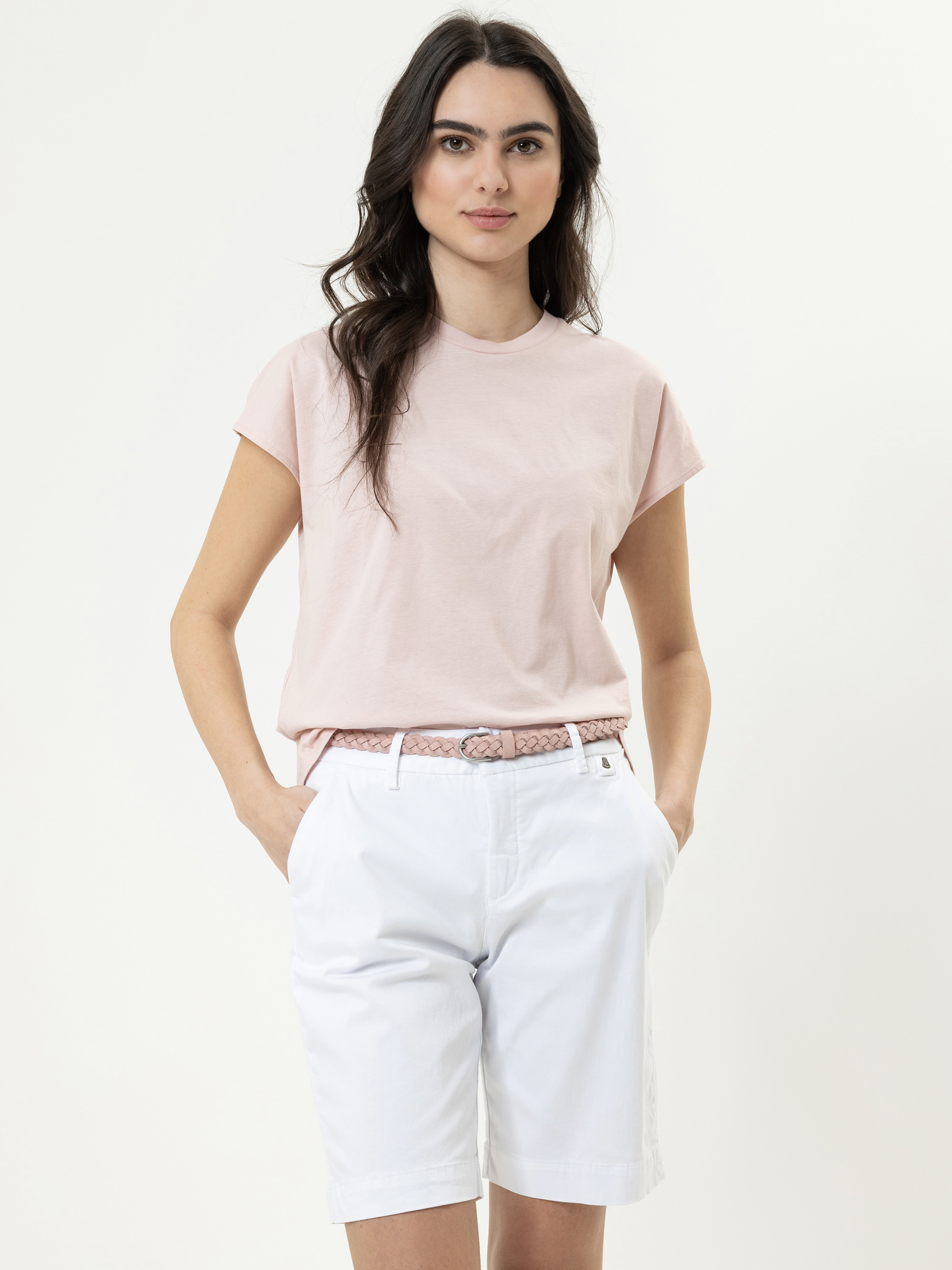 Eine Frau mit langen dunklen Haaren, die ein blassrosa T-Shirt, weiße knielange Shorts und einen geflochtenen rosa Gürtel trägt, steht mit den Händen in den Taschen vor einem schlichten hellen Hintergrund.