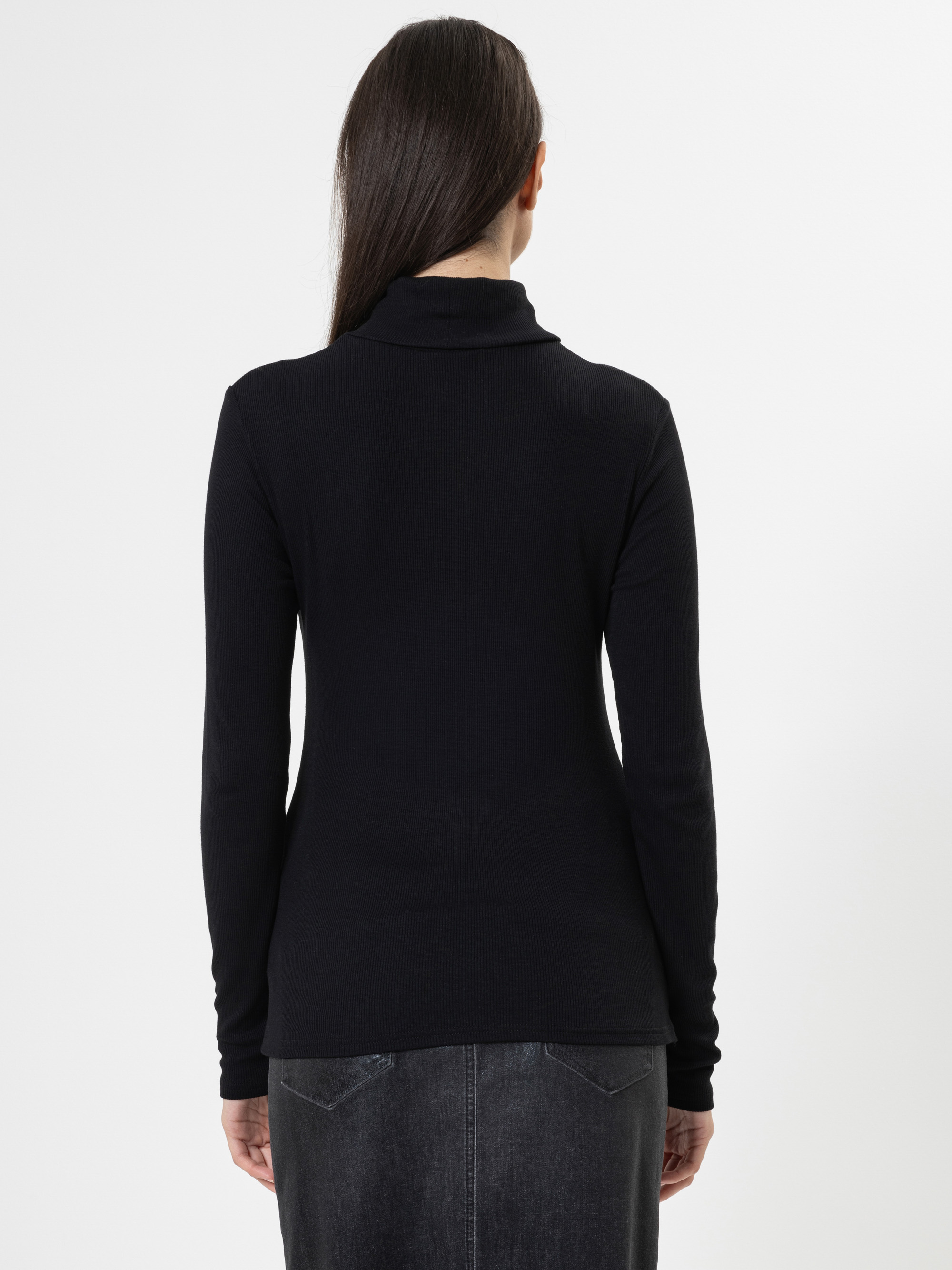 Eine Person mit langen dunklen Haaren wird von hinten gezeigt, die einen schwarzen langärmeligen Rollkragenpullover und einen dunkelgrauen Rock vor einem schlichten weißen Hintergrund trägt.