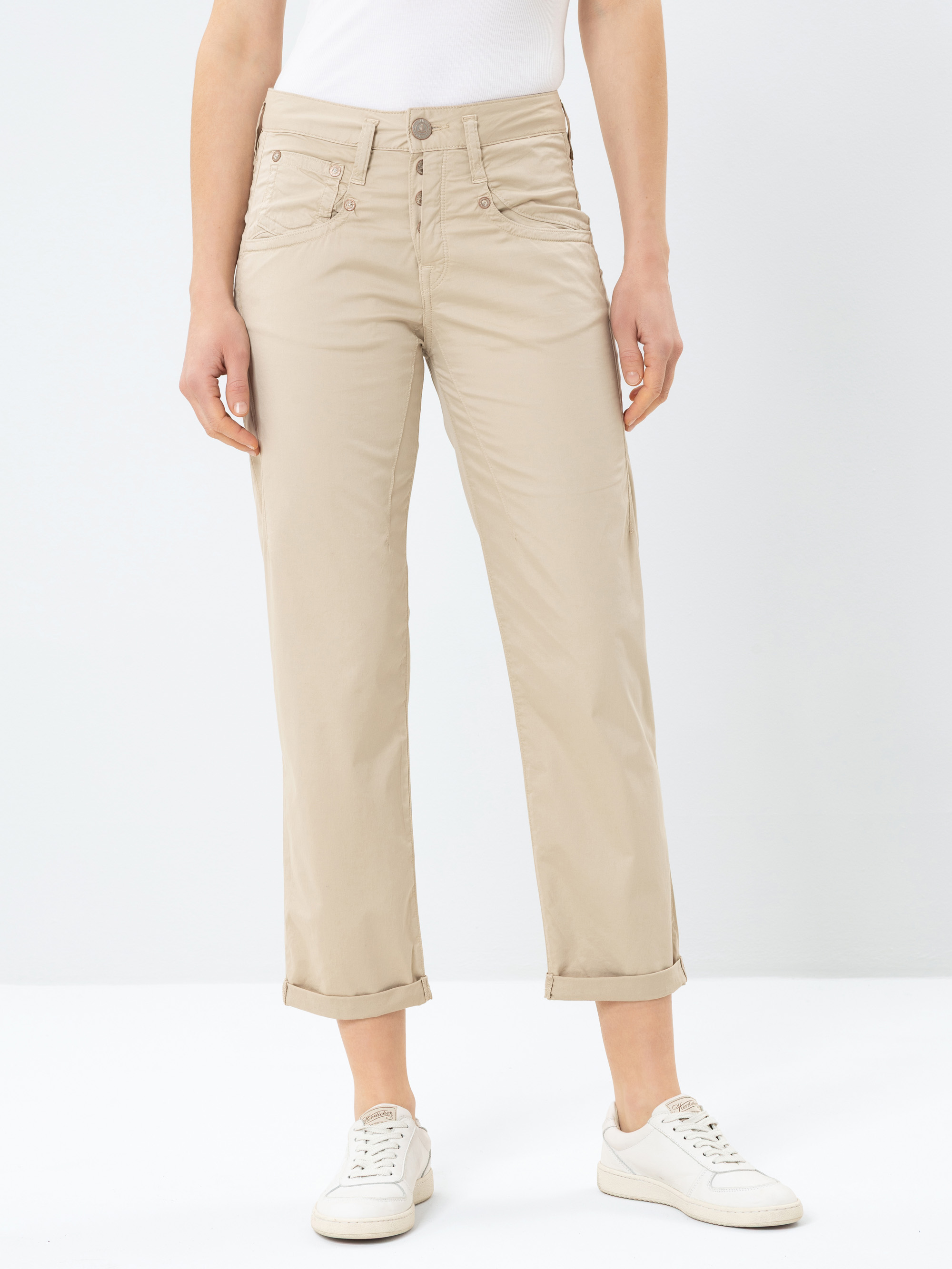 Person, die eine hellbeige Hose mit geradem Bein und Bündchen, weiße Turnschuhe und ein weißes Oberteil trägt und vor einem schlichten weißen Hintergrund steht. Der Kopf der Person ist auf dem Bild nicht zu sehen.