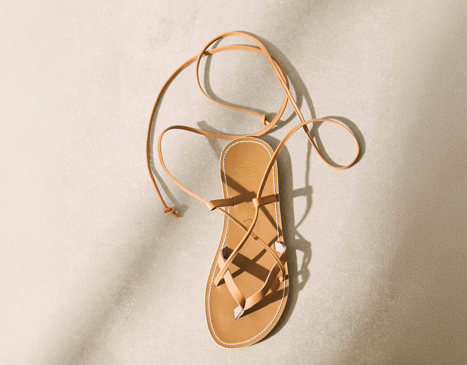 Eine einzelne hellbraune Ledersandale mit dünnen Riemen und einer langen Spitze, die auf einer hellen, sandigen Oberfläche mit weichen Schatten liegt.