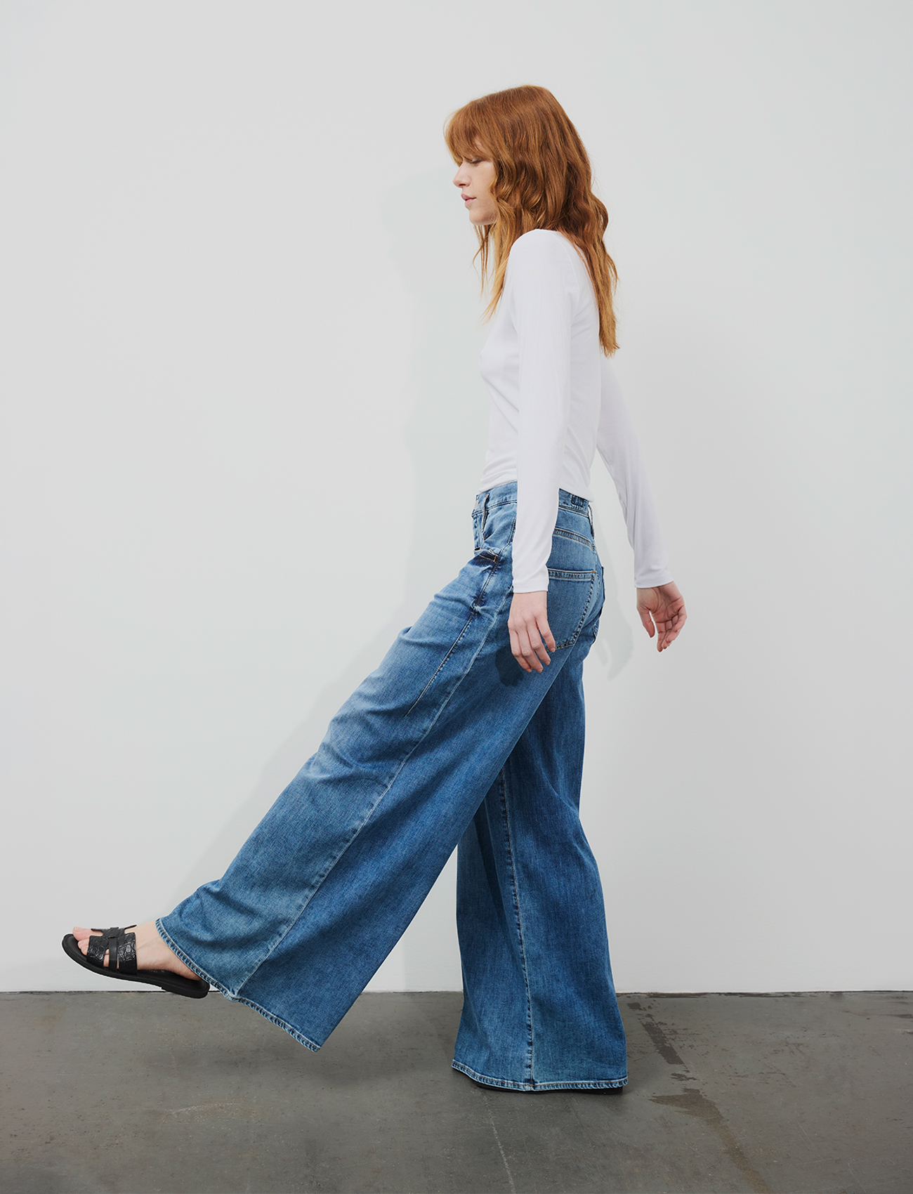 Eine Person mit langen roten Haaren, die ein weißes Langarmshirt, eine blaue Jeans mit weitem Bein und schwarze Sandalen trägt, steht im Profil vor einer schlichten weißen Wand und einem grauen Boden und hebt einen Fuß, als würde sie einen Schritt ma