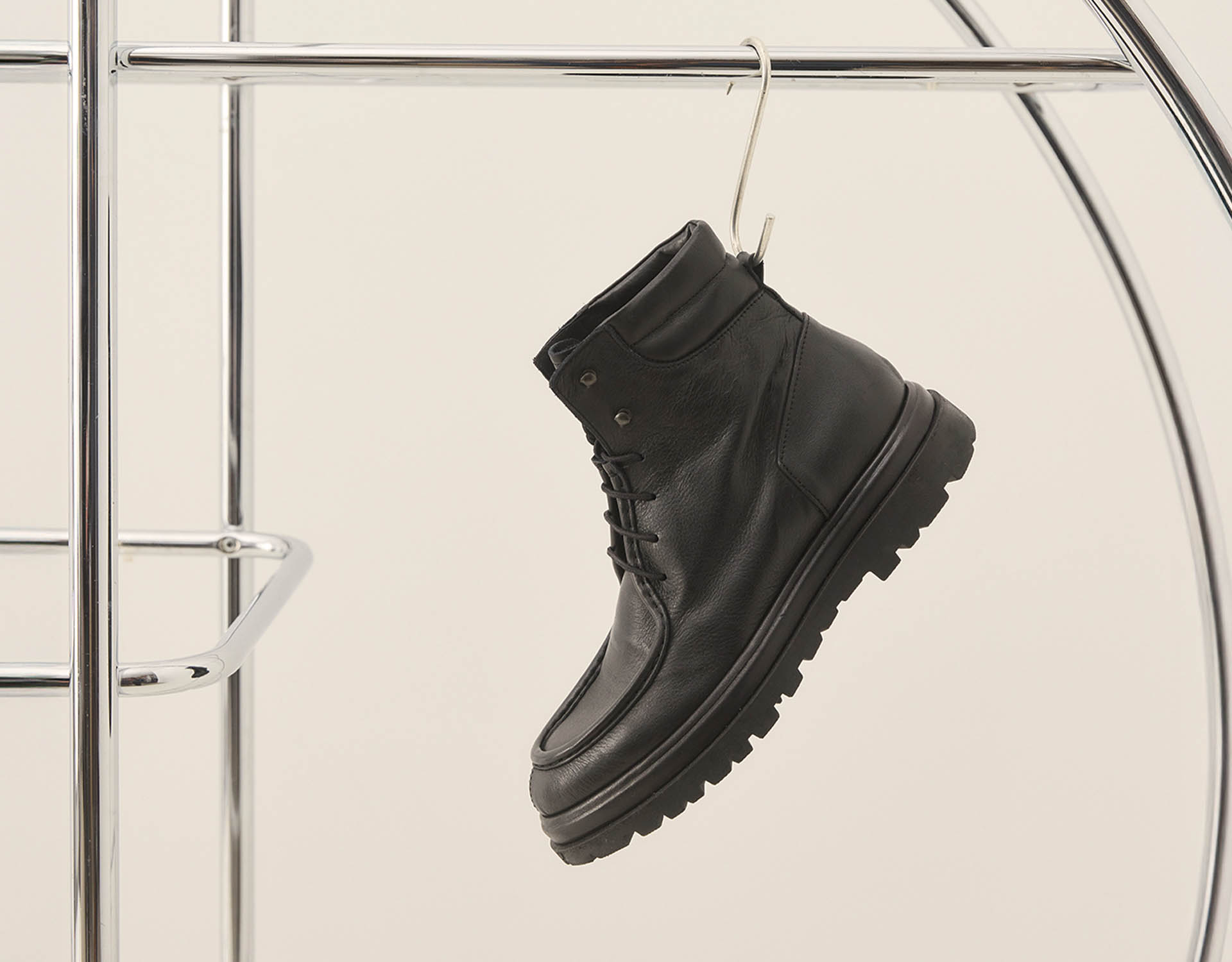 Ein schwarzer Schnürstiefel aus Leder mit einer dicken Sohle hängt an einem Metallhaken an einem polierten Metallkleiderständer vor einem schlichten, hellen Hintergrund.