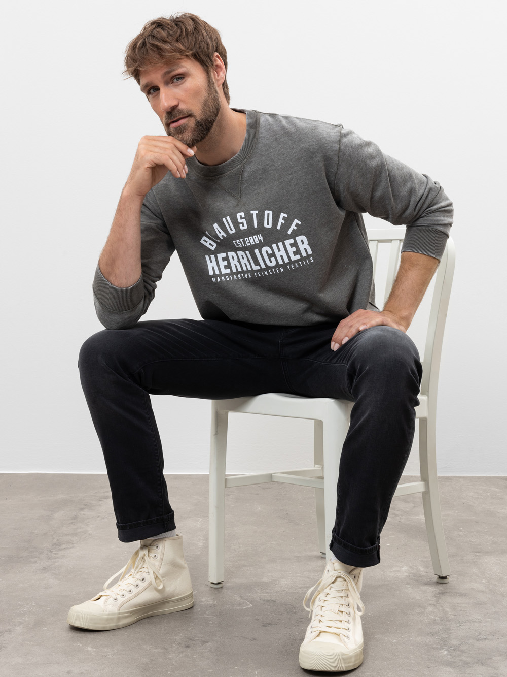 Ein Mann mit hellbraunem Haar und Bart sitzt auf einem weißen Stuhl, trägt ein graues Sweatshirt mit weißer Aufschrift, schwarze Jeans und weiße High-Top-Turnschuhe und posiert mit einer Hand an seinem Kinn.