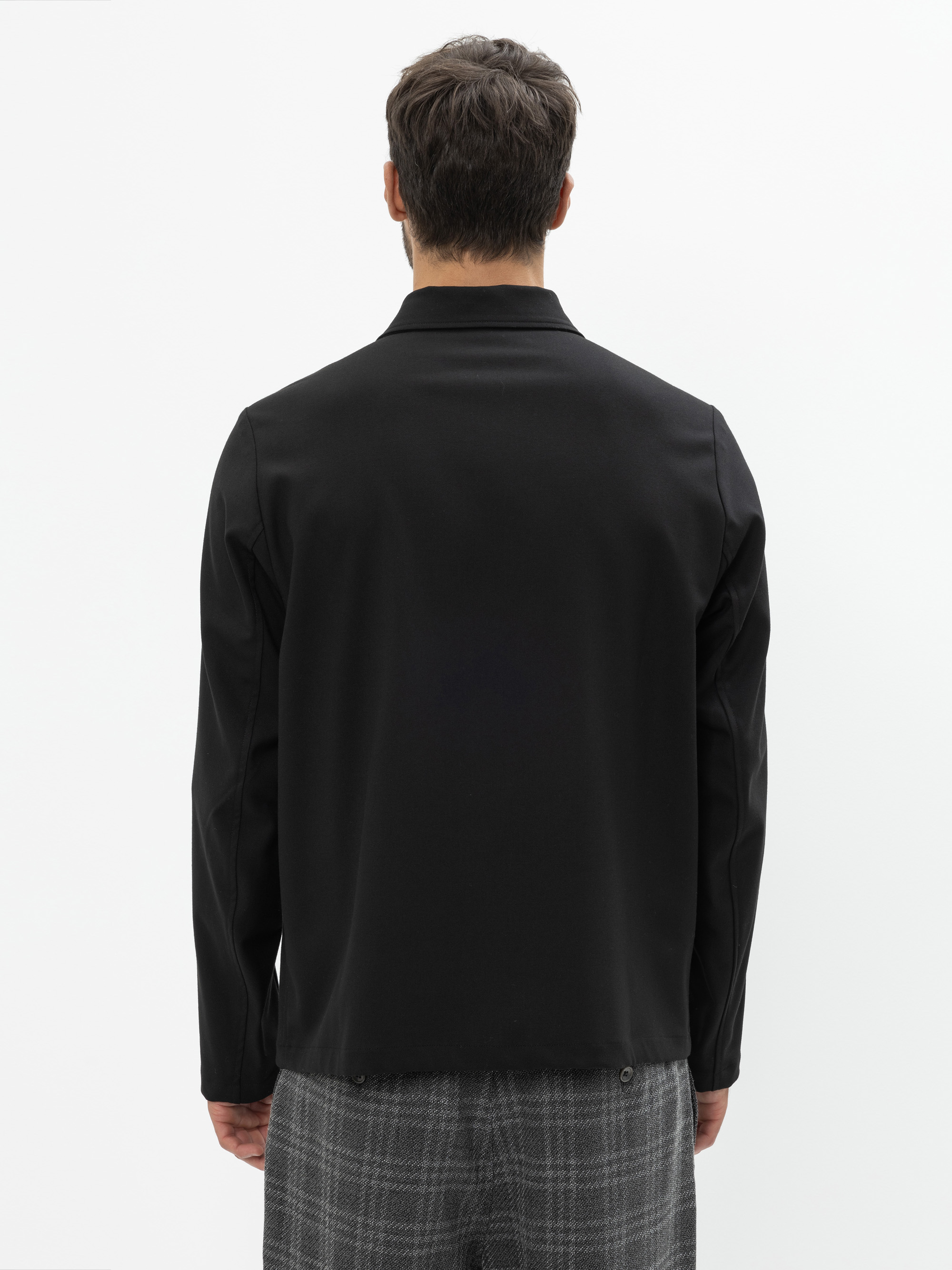 Ein Mann steht mit dem Rücken zur Kamera, trägt ein schwarzes langärmeliges Hemd und eine grau karierte Hose vor einem schlichten weißen Hintergrund.