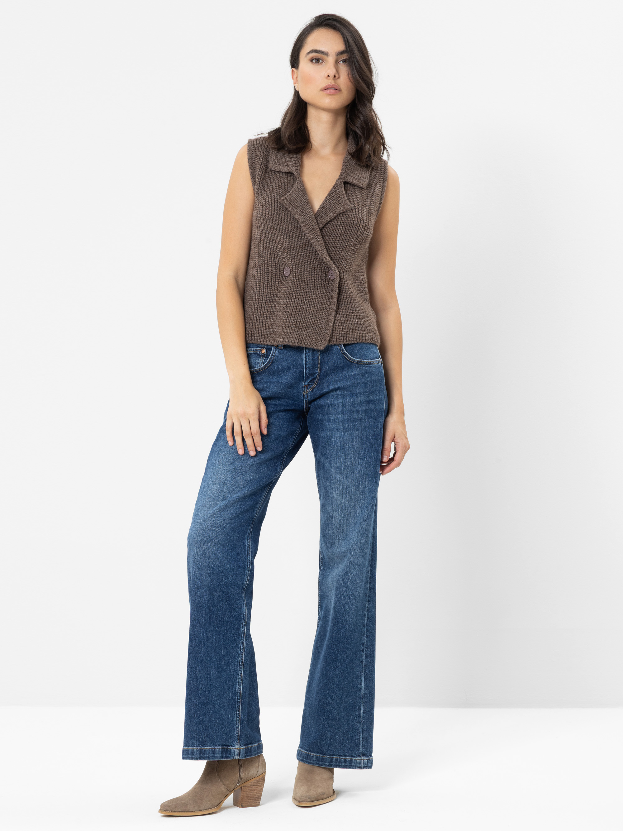 Eine Frau steht vor einem schlichten weißen Hintergrund. Sie trägt eine braune, ärmellose Strickweste, eine blaue Jeans mit weitem Bein und beigefarbene Stiefeletten; ihr Haar fällt ihr offen ins Gesicht, und ihr Gesichtsausdruck ist neutral.