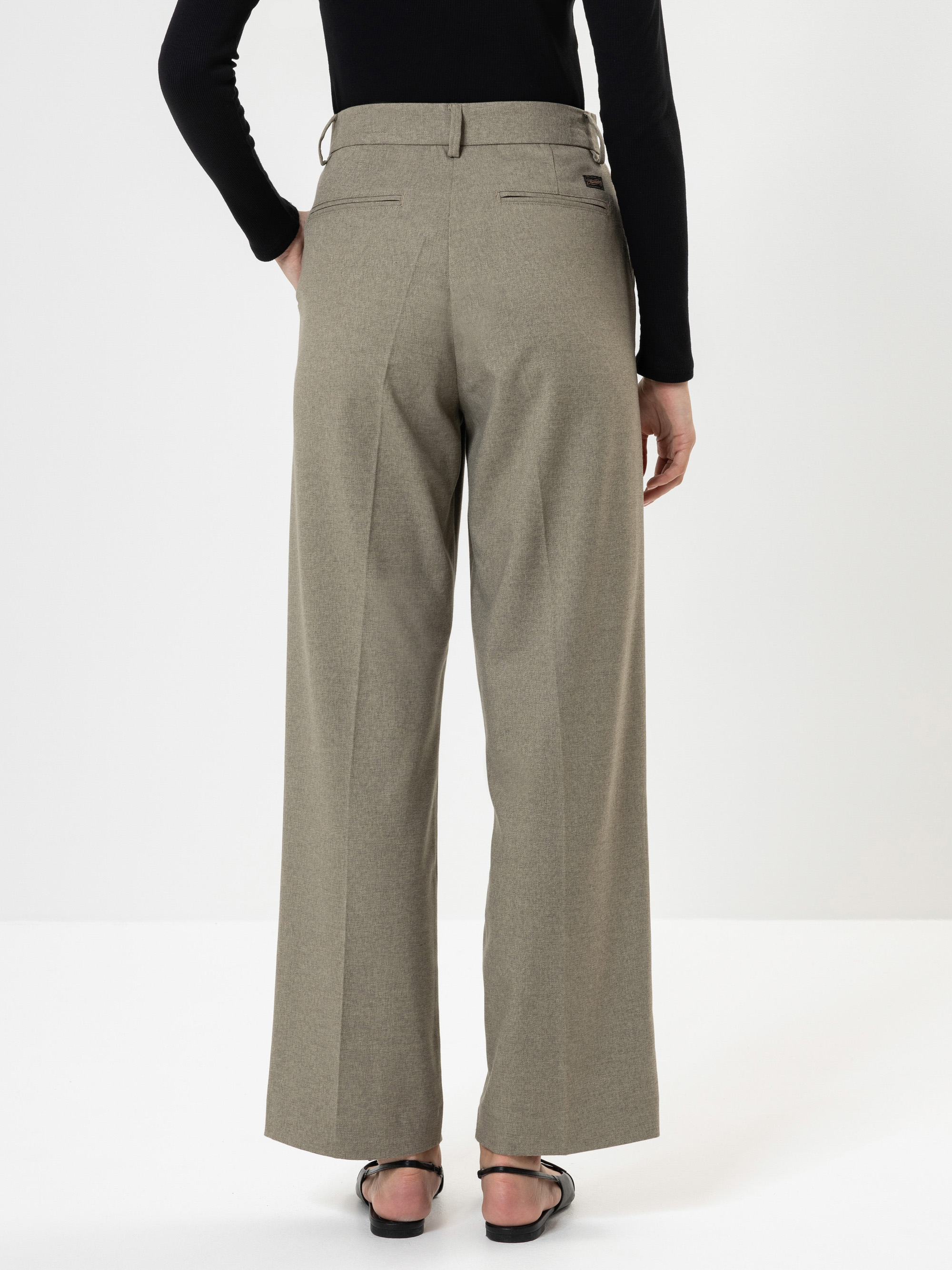 Eine Person, die ein schwarzes langärmeliges Oberteil, eine beigefarbene Hose mit weitem Bein und schwarze Sandalen trägt, steht mit dem Rücken zur Kamera vor einem schlichten weißen Hintergrund.