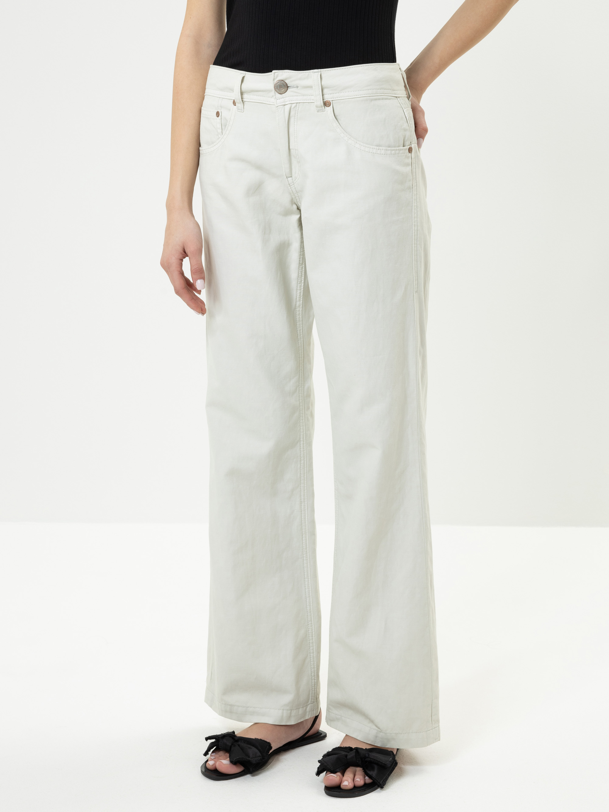 Eine Person, die eine cremefarbene Jeans mit weitem Bein und ein schwarzes ärmelloses Oberteil trägt, steht vor einem schlichten weißen Hintergrund. Ihre Füße stecken in schwarzen Sandalen mit Schleifendetails. Ihr Kopf ist auf dem Bild nicht zu sehe