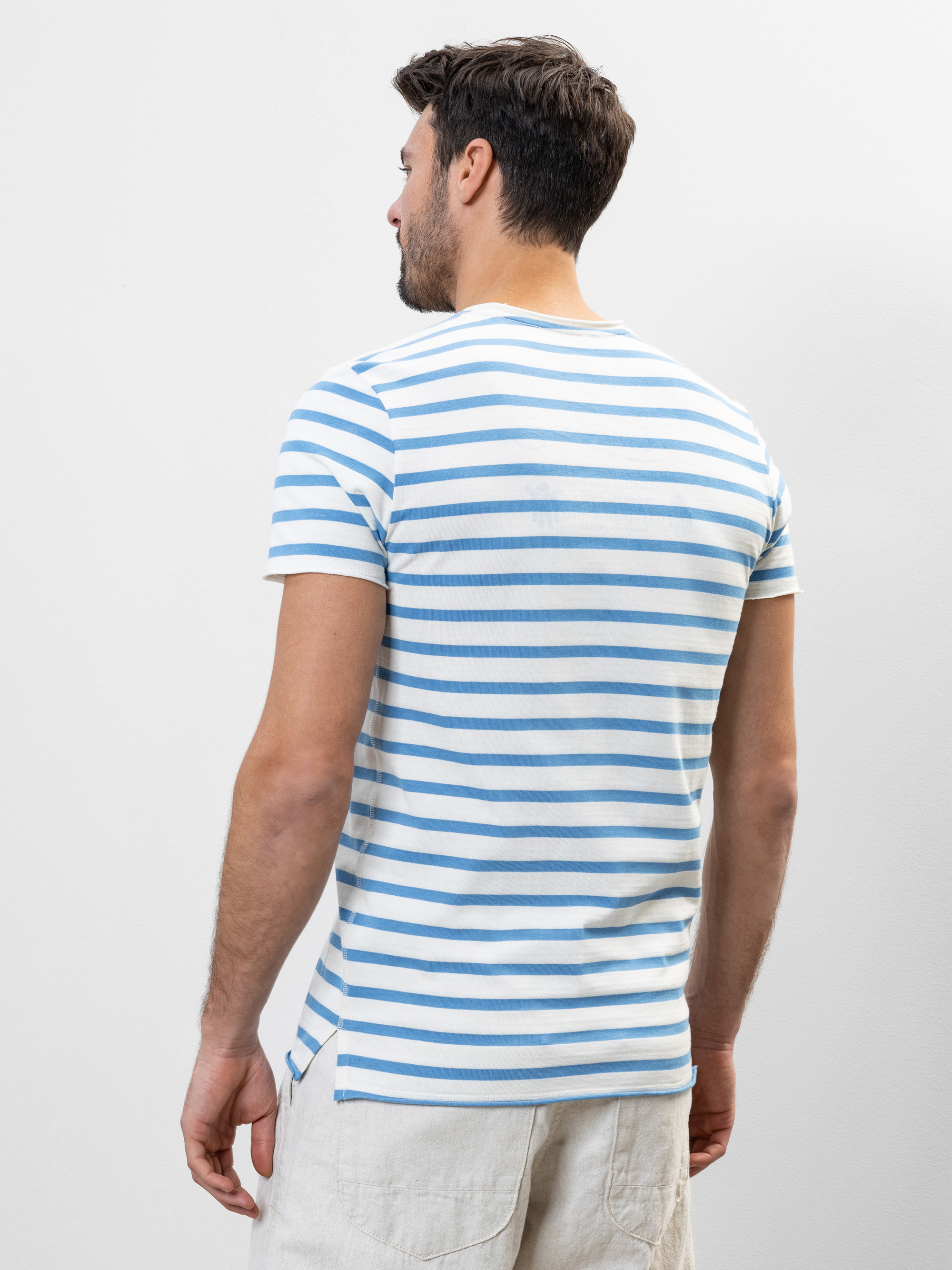 Ein Mann mit kurzen braunen Haaren steht mit dem Rücken zur Kamera, trägt ein weißes T-Shirt mit blauen Querstreifen und helle Shorts vor einem schlichten hellen Hintergrund.