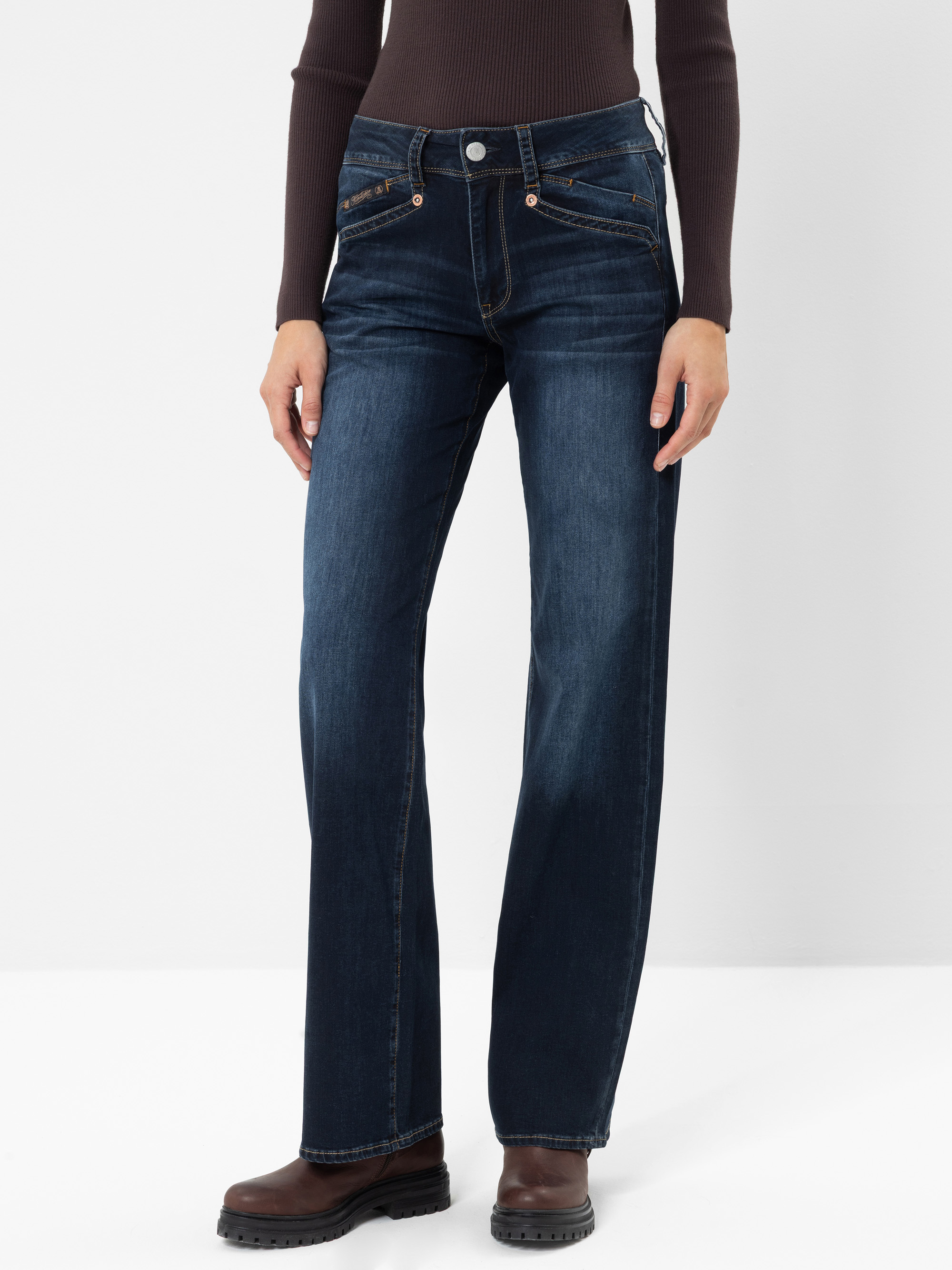 Eine Person, die ein dunkelbraunes Langarmshirt, eine dunkelblaue Bootcut-Jeans mit Kontrastnähten und braune Stiefel trägt und vor einem weißen Hintergrund steht.