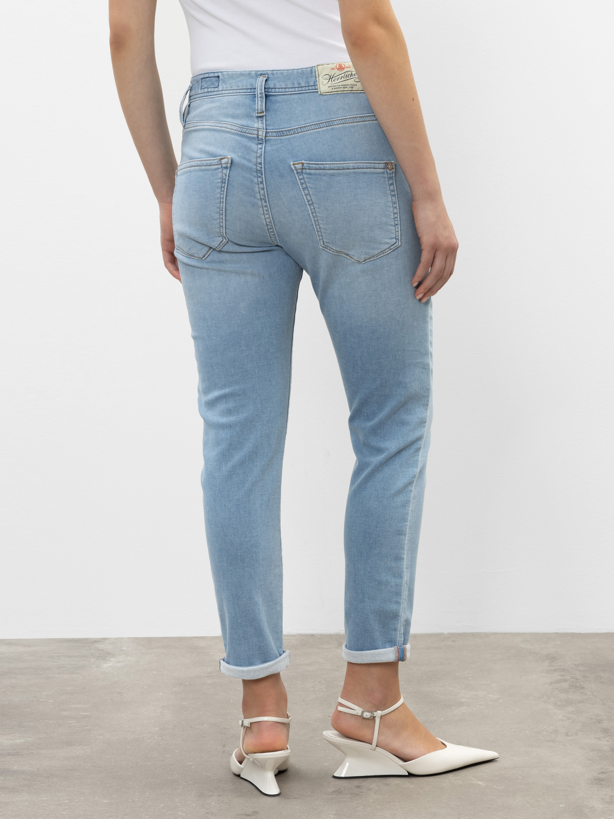 Eine Person, die hellblaue Jeans mit Bündchen, ein weißes Oberteil und weiße, spitz zulaufende Halbschuhe trägt, steht auf einem grauen Boden und blickt von der Kamera weg.