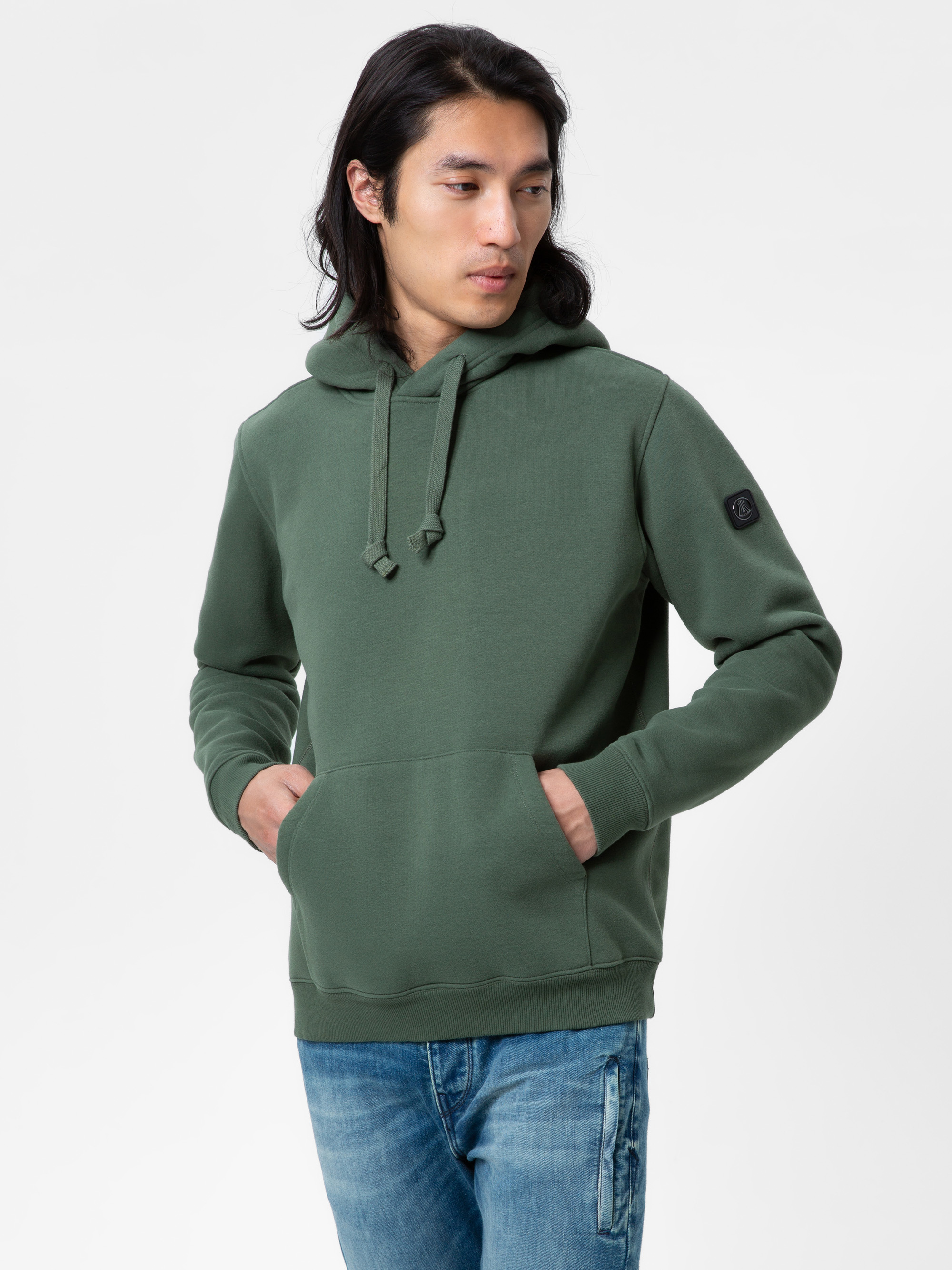 Eine Person mit langen dunklen Haaren trägt einen grünen Kapuzenpulli mit einer Vordertasche und hellblaue Jeans. Sie steht vor einem schlichten weißen Hintergrund und blickt nach links.