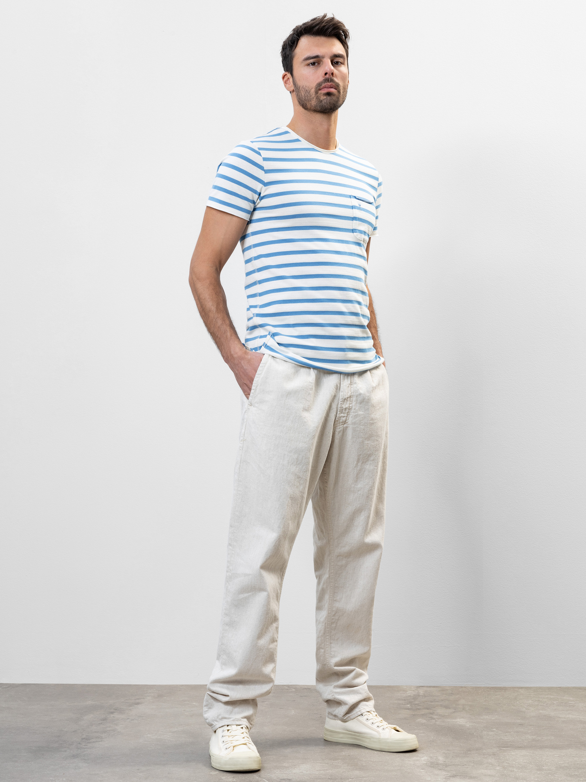 Ein Mann steht vor einem schlichten Hintergrund. Er trägt ein hellblau-weiß gestreiftes T-Shirt, eine wollweiße, weite Hose und weiße Turnschuhe. Er hat kurzes dunkles Haar und einen gestutzten Bart, die Hände stecken in den Taschen.