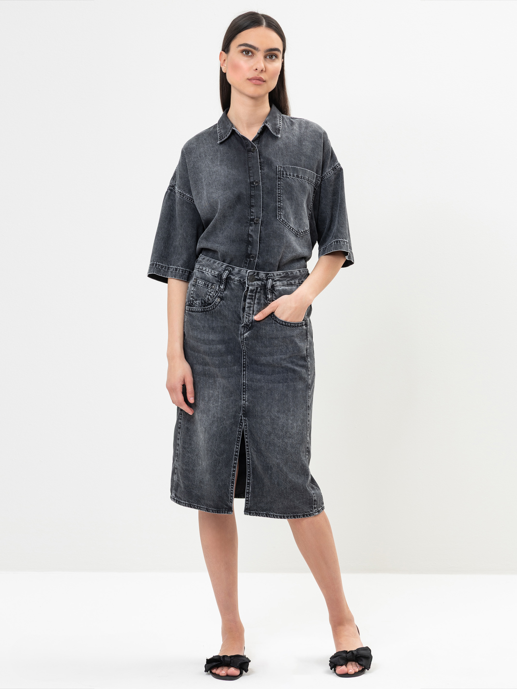 Eine Frau steht vor einem schlichten weißen Hintergrund und trägt ein verblichenes schwarzes Jeanshemd mit gekrempelten Ärmeln und einen passenden knielangen Jeansrock. Sie hat eine Hand in der Rocktasche und trägt schwarze, zehenoffene Sandalen.