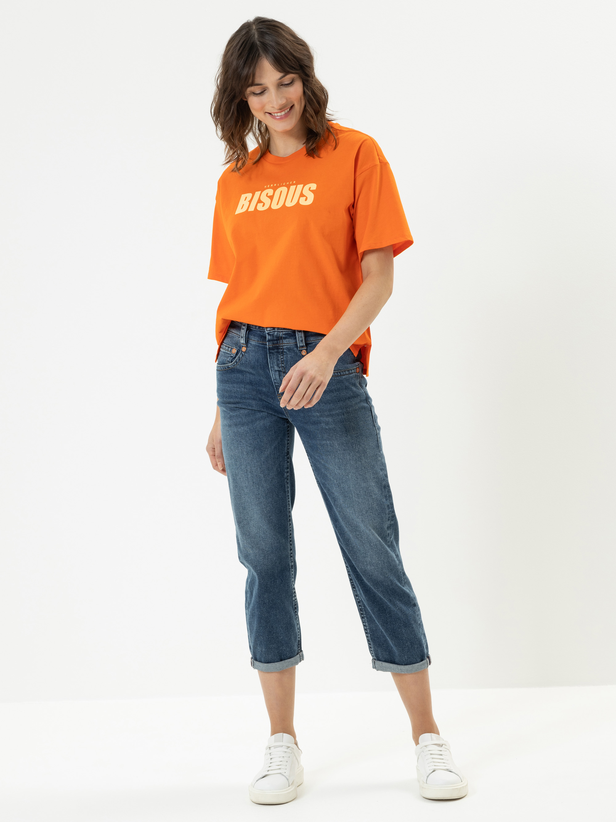 Eine Frau steht vor einem schlichten Hintergrund und trägt ein orangefarbenes T-Shirt mit dem Aufdruck BISOUS, blaue Jeans und weiße Turnschuhe. Sie lächelt und schaut nach unten.
