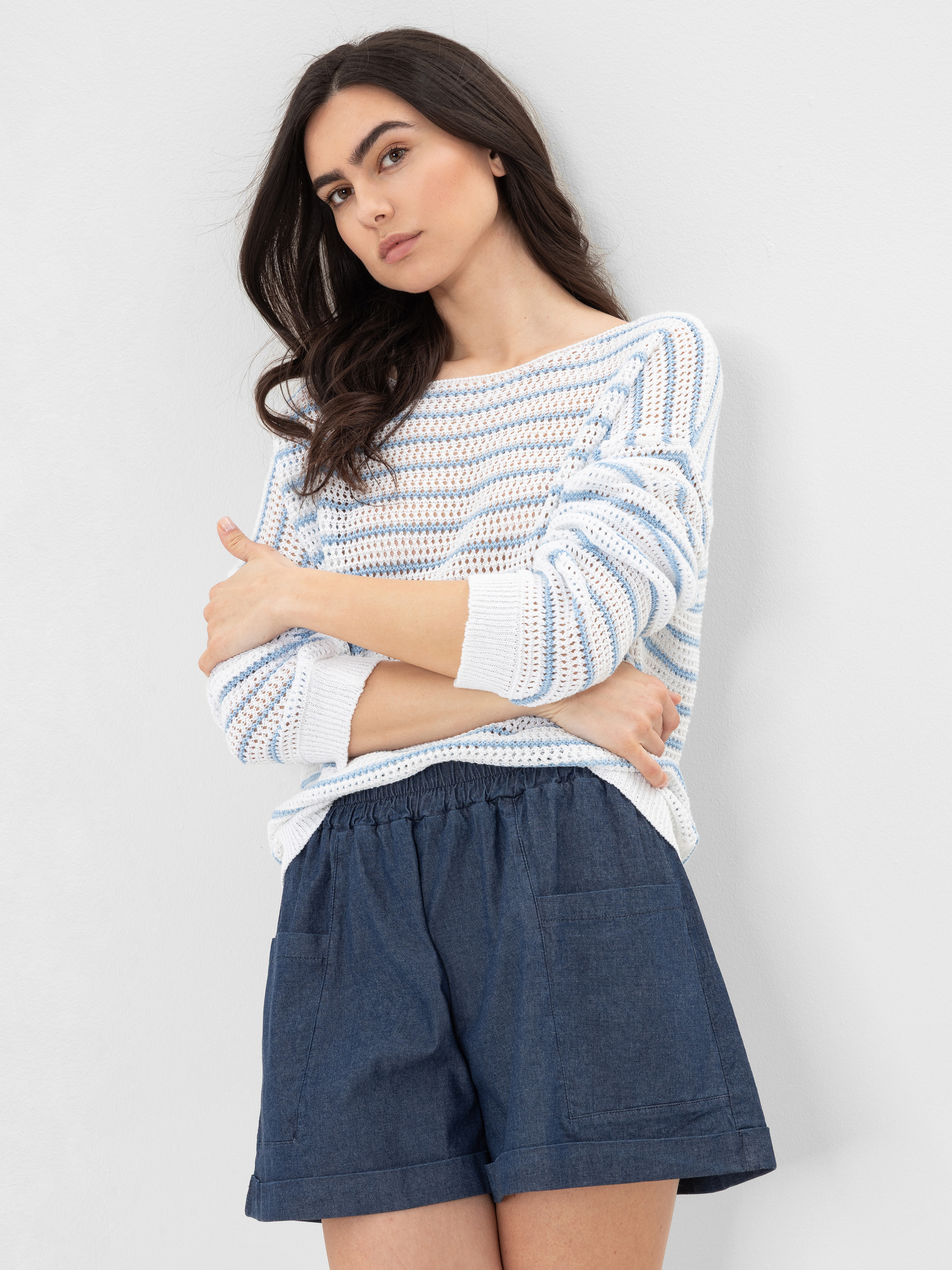 Eine Frau mit langen dunklen Haaren steht vor einer weißen Wand. Sie trägt einen lockeren weiß-blau gestreiften Strickpullover und blaue Shorts mit hoher Taille und Vordertaschen. Sie hat die Arme verschränkt und einen neutralen Gesichtsausdruck.