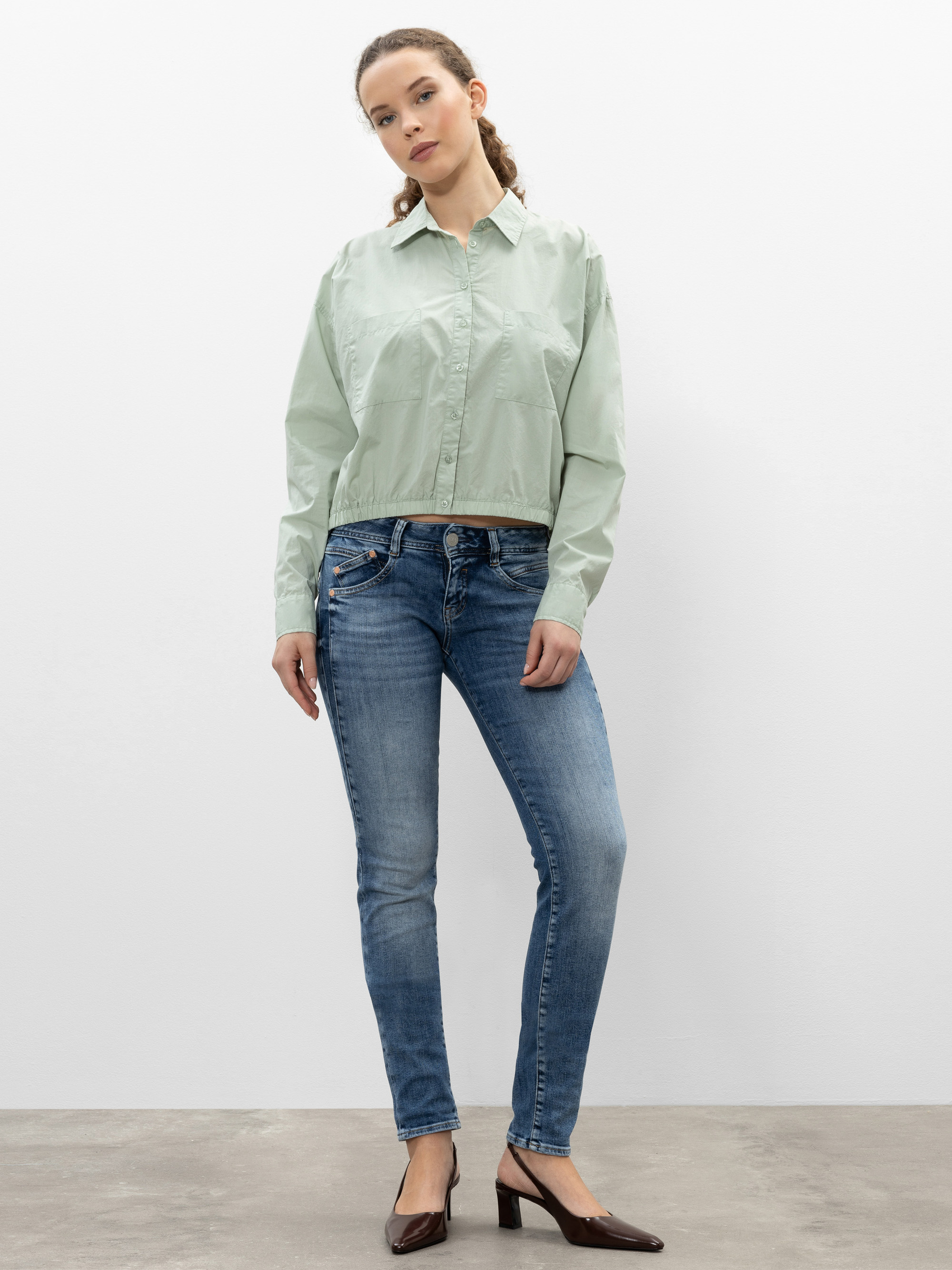 Eine Frau steht vor einer schlichten weißen Wand und trägt ein hellgrünes Button-up-Hemd, blaue Jeans und dunkelbraune Halbschuhe. Sie hat ihr Haar zurückgebunden und blickt ruhig in die Kamera.