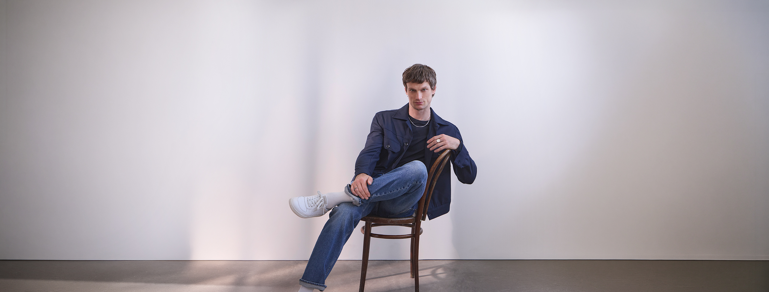 Eine Person mit kurzen Haaren sitzt auf einem Holzstuhl in einem minimalistisch eingerichteten, gut beleuchteten Raum. Sie trägt eine marineblaue Jacke, ein dunkles Hemd, blaue Jeans und weiße Sneaker, hat ein Bein über das andere geschlagen und zeigt