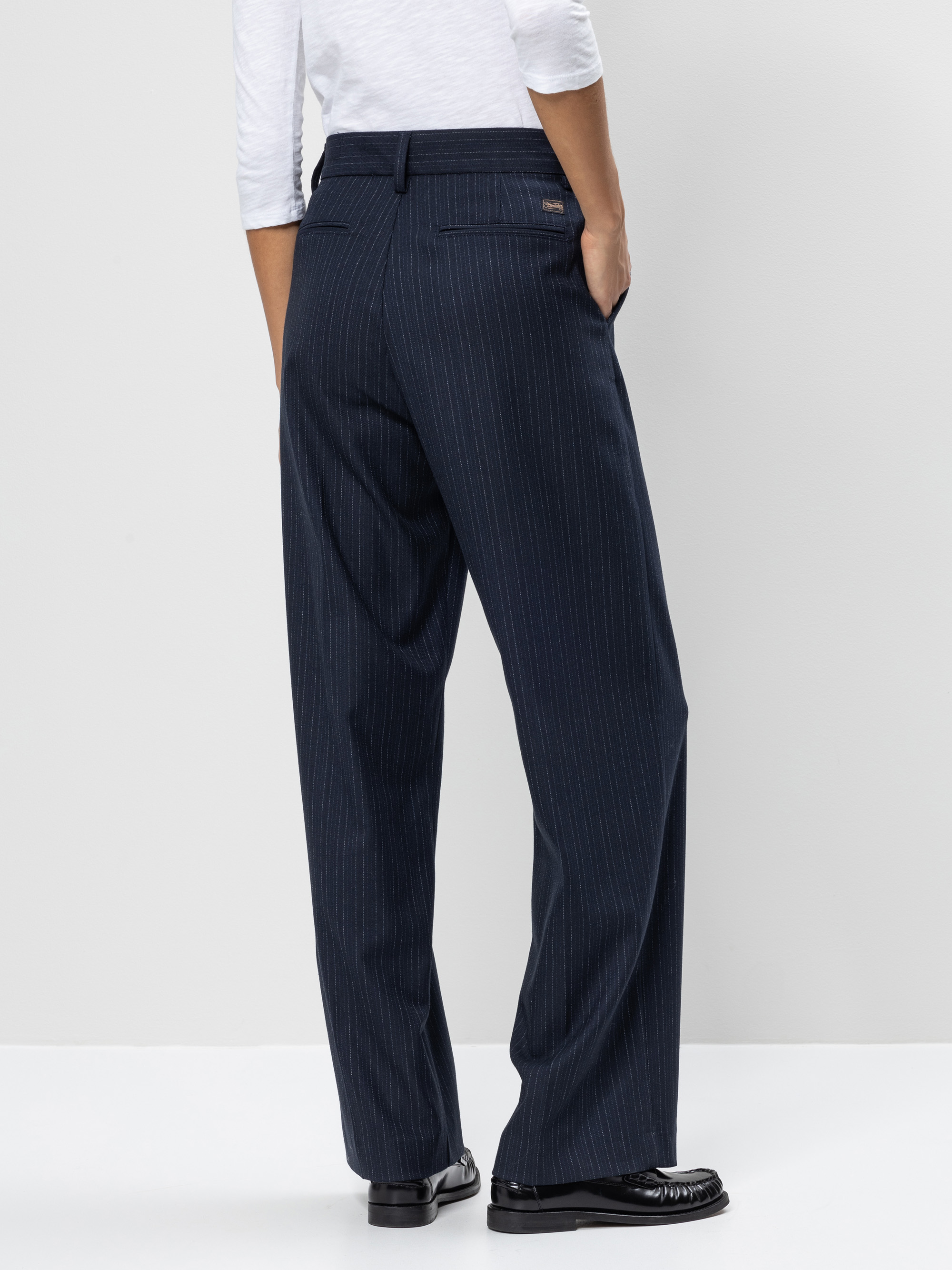 Eine Person steht mit dem Gesicht zur Wand, trägt ein weißes langärmeliges Hemd und eine dunkelblaue Hose mit weitem Bein in Nadelstreifen und schwarzen Halbschuhen vor einem schlichten hellen Hintergrund.