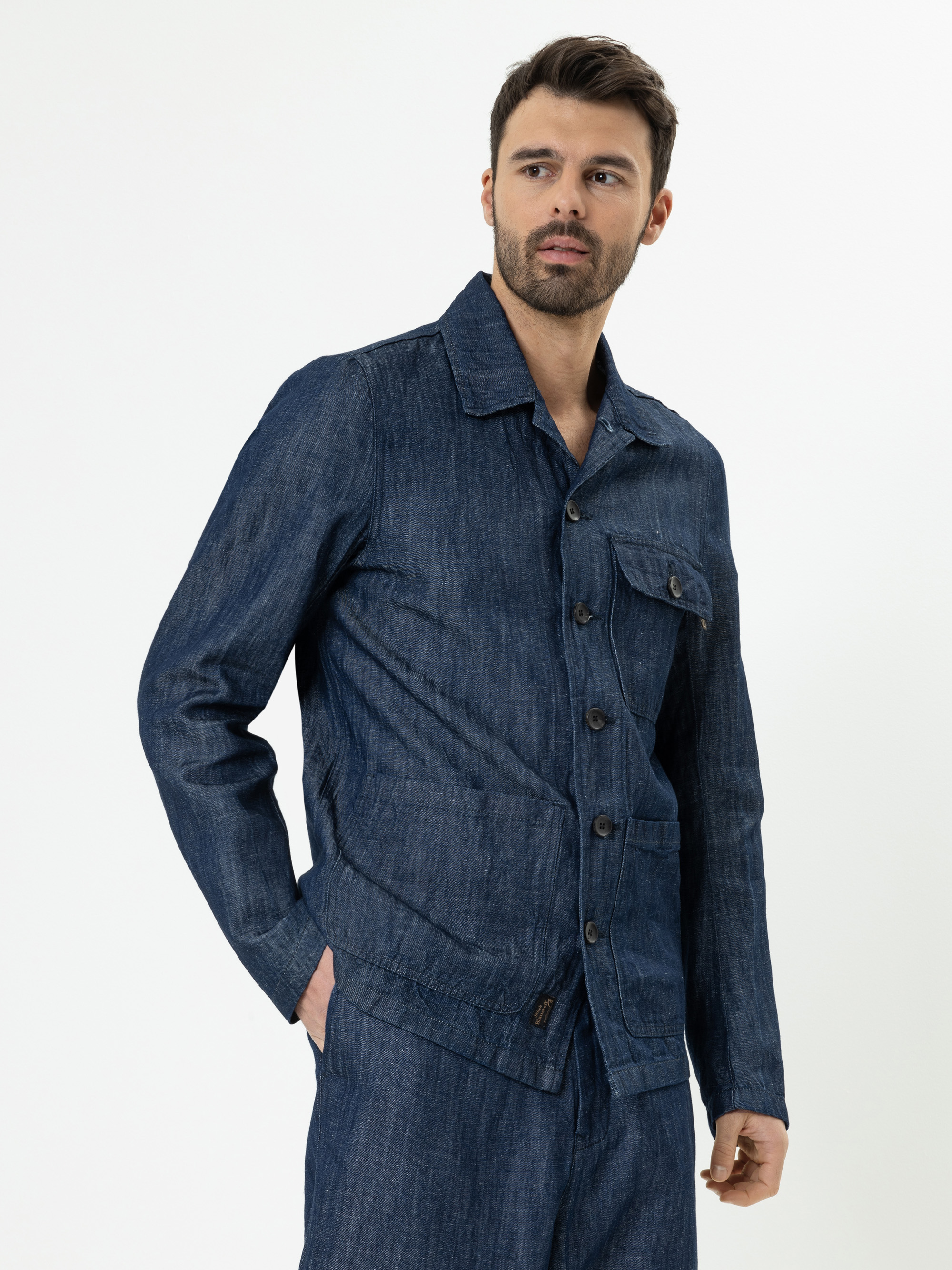 Ein Mann mit kurzen dunklen Haaren und gestutztem Bart trägt eine blaue Jeansjacke und eine passende Hose und steht mit einer Hand in der Tasche vor einem schlichten weißen Hintergrund.